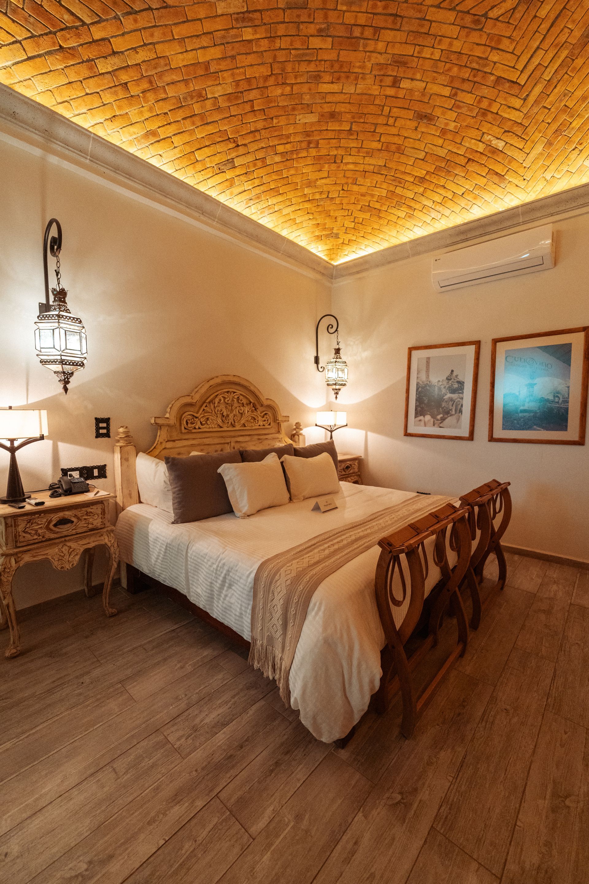 Dormitorio con cama de madera ornamentada, techo de ladrillo, lámparas colgantes y obras de arte enmarcadas.
