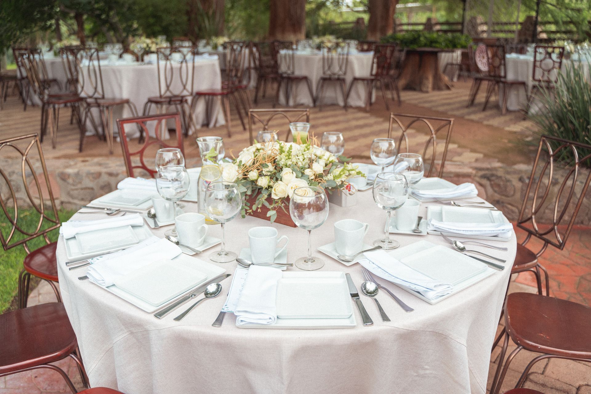Outdoor wedding reception: round tables with white linens, floral centerpieces, set for guests.
