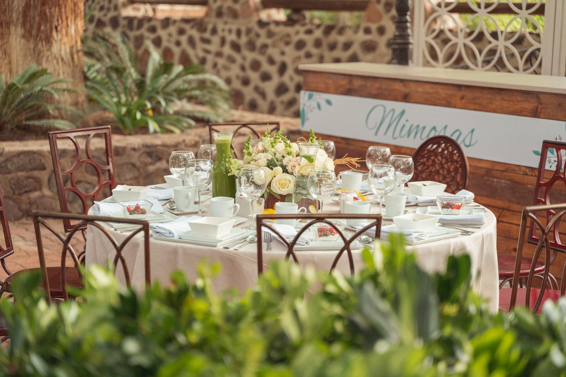 Outdoor table set for a meal, with floral arrangement, glasses, and chairs, likely a brunch.