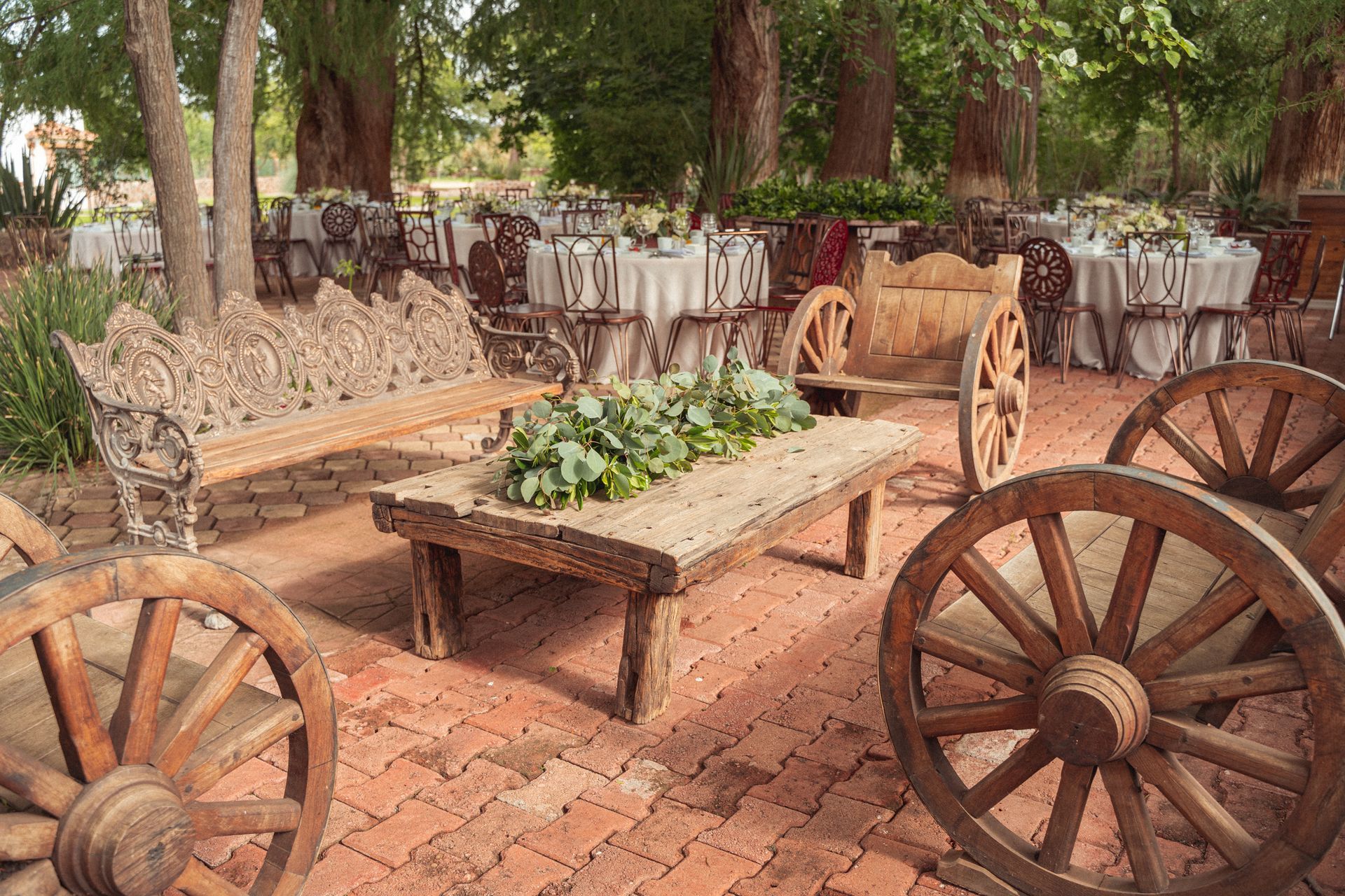 Outdoor dining area with tables set for a gathering, featuring rustic wooden benches, wagon wheels, and a brick patio.