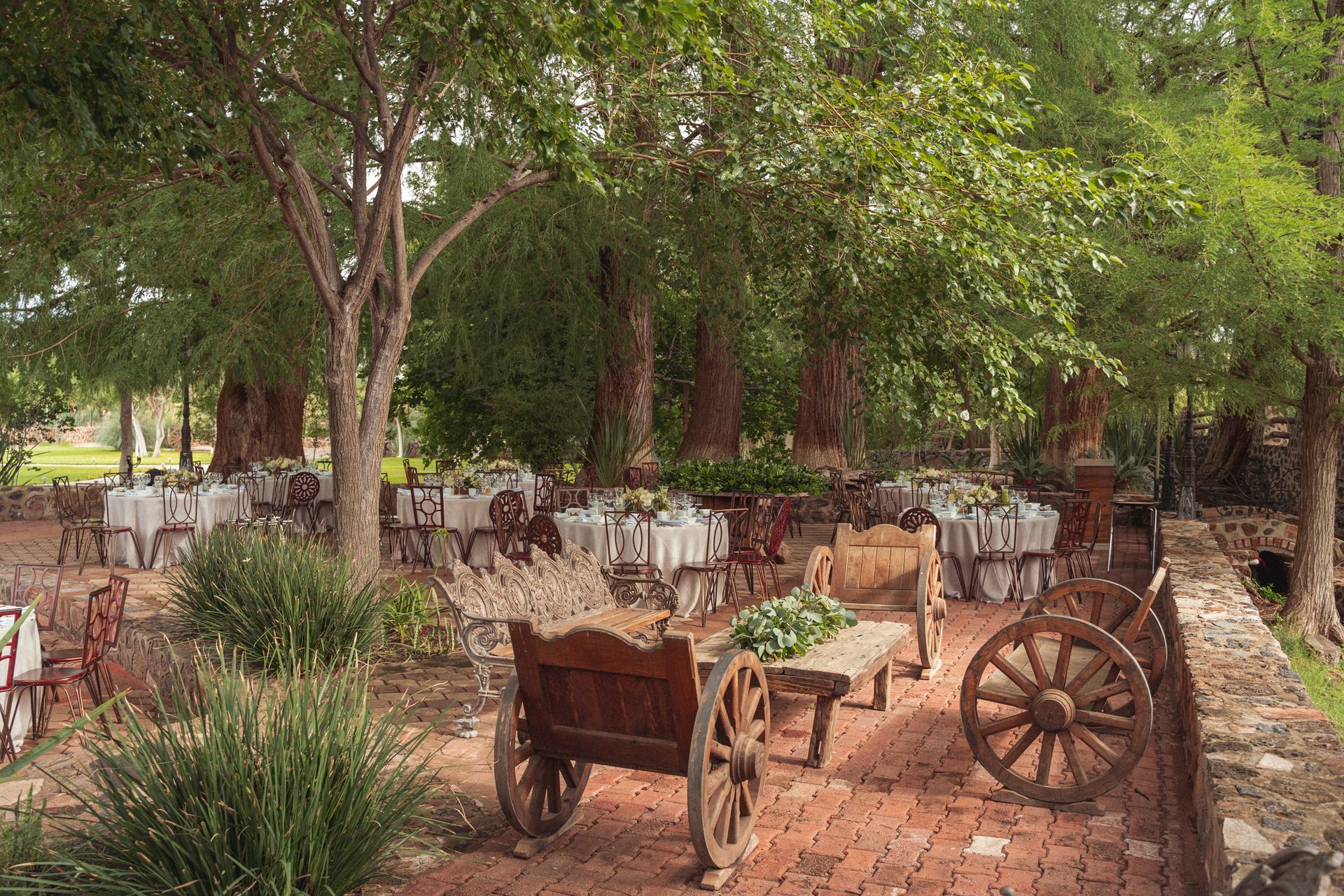 Outdoor dining tables set under trees, with wooden wagons on a brick patio.