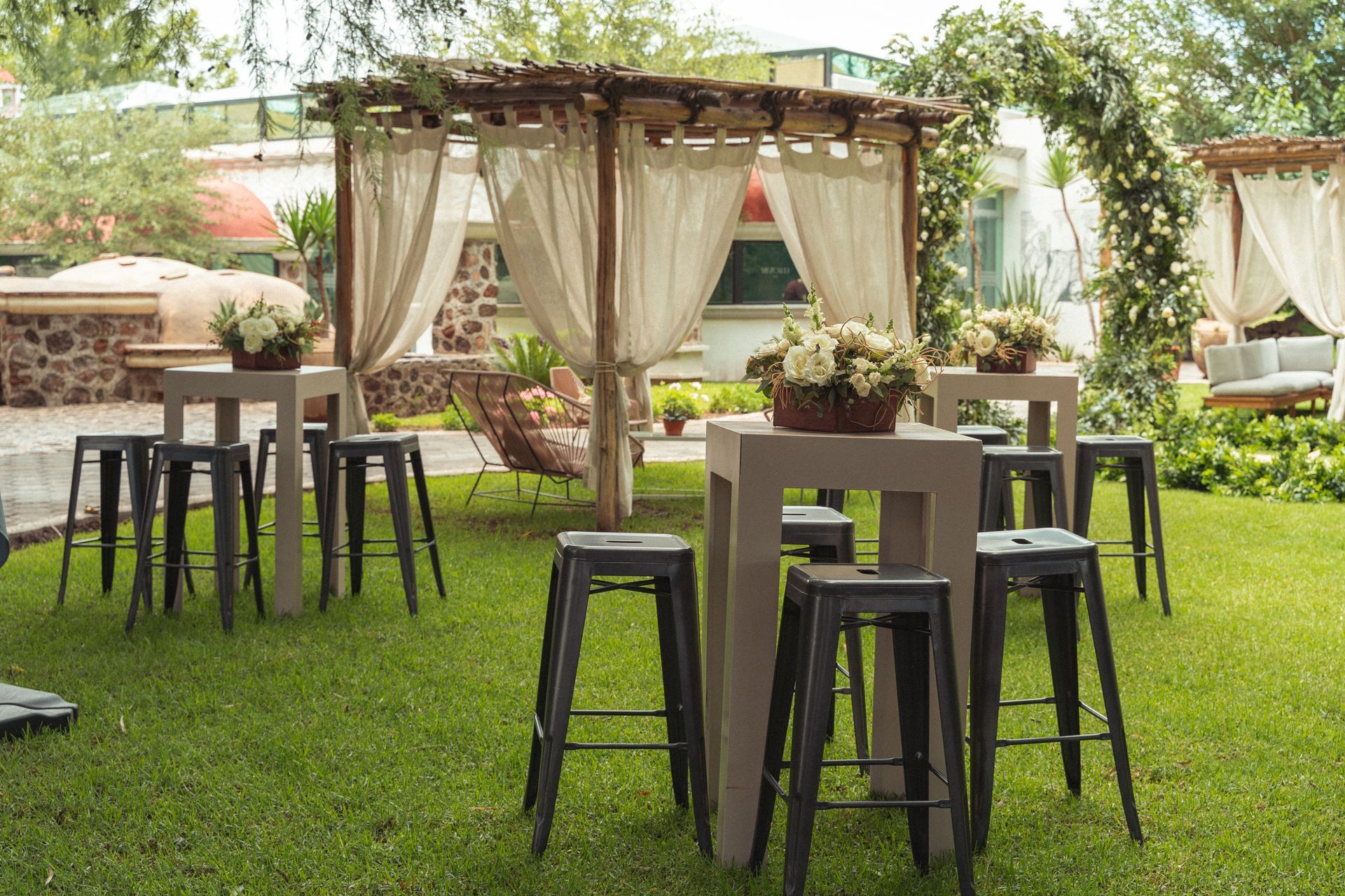 Outdoor event with high tables, black stools, floral centerpieces, and draped pergolas on a lawn.