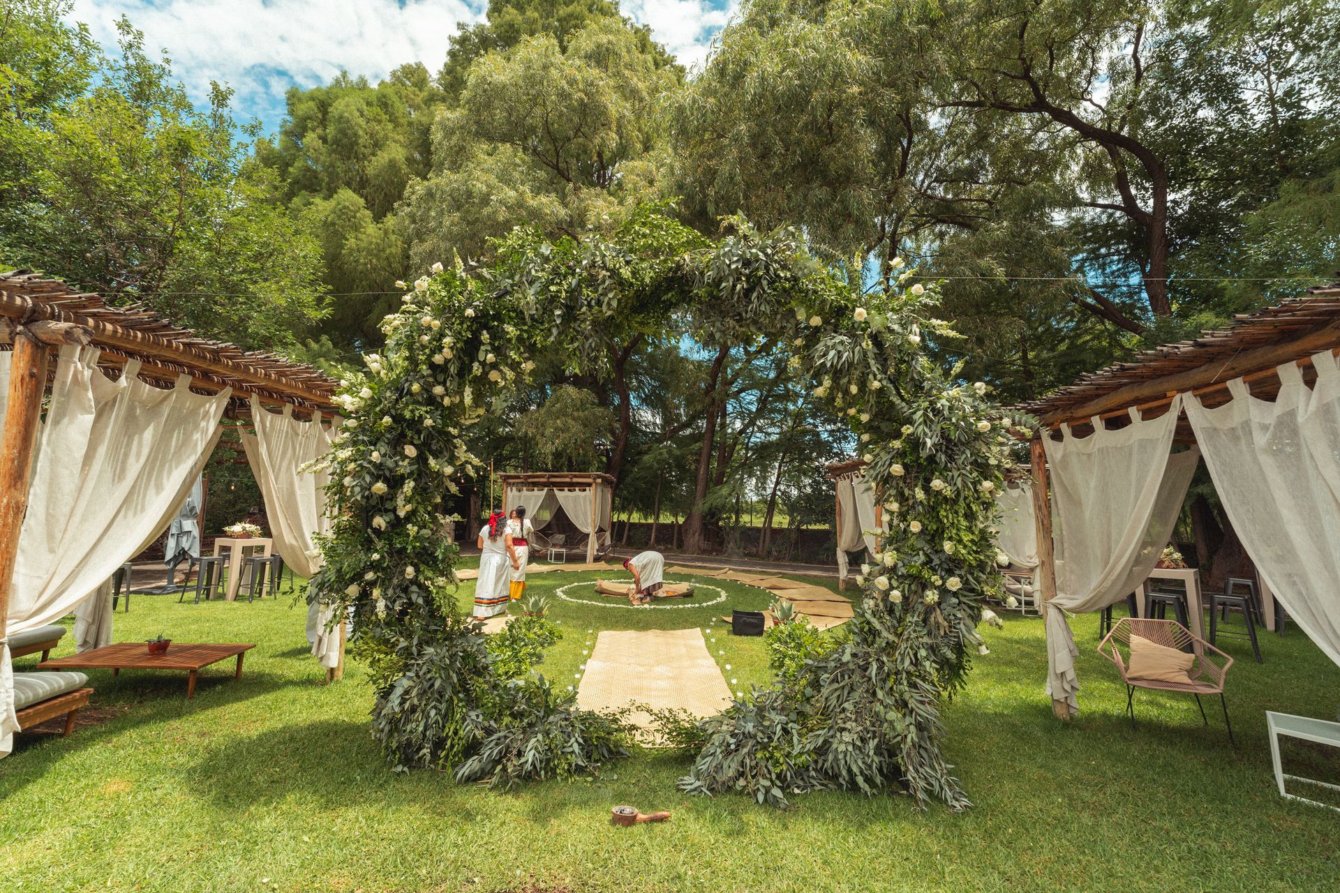 Wedding arch with floral decorations on a grassy lawn; white canopies and trees in the background.