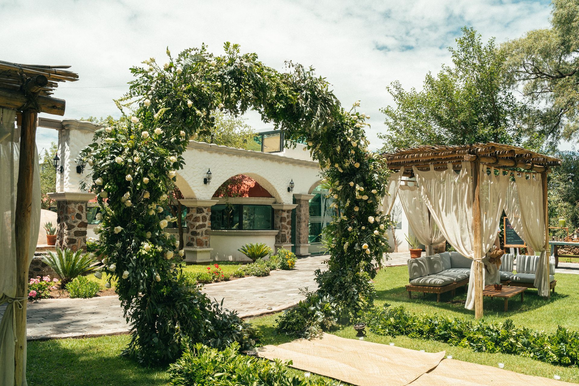 Wedding ceremony archway covered in greenery, leading to a draped gazebo in a garden setting.