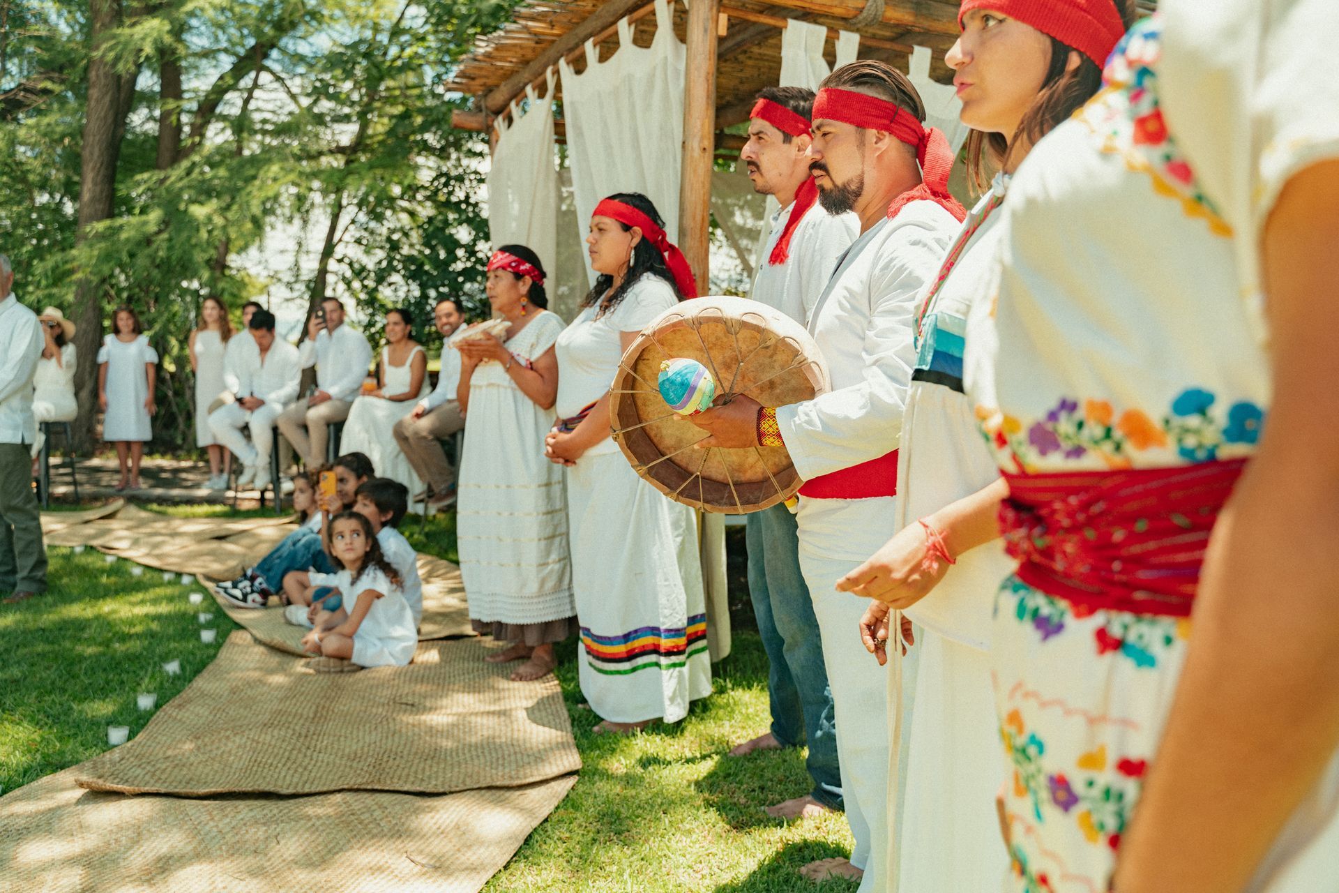 Personas vestidas de blanco con cintas rojas en la cabeza; una ceremonia al aire libre; una persona sostiene un tambor.