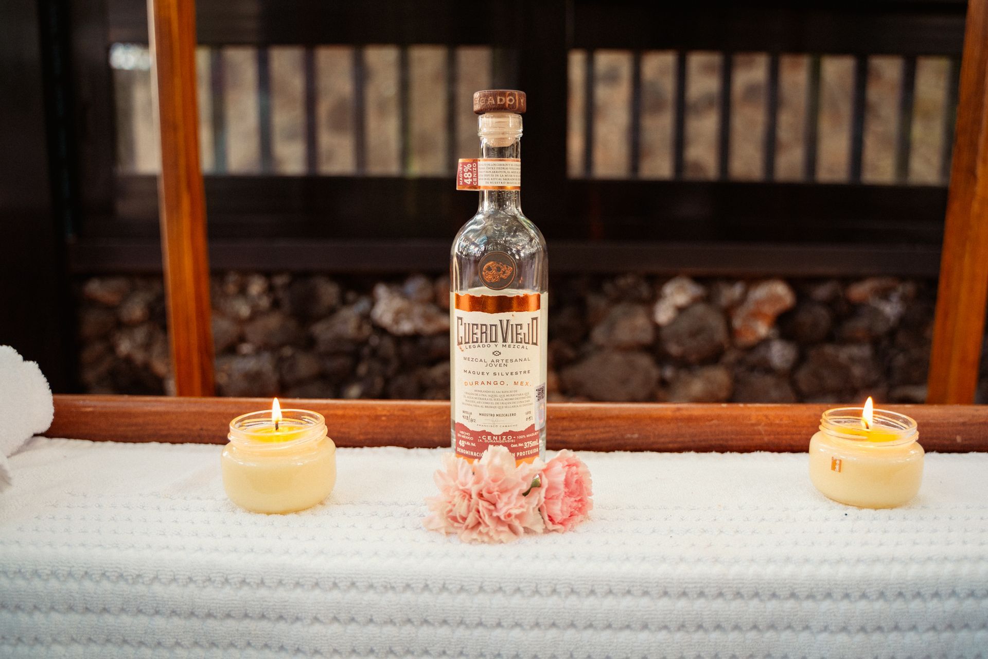 Botella de licor, flores y velas sobre una superficie blanca, con fondo de madera.