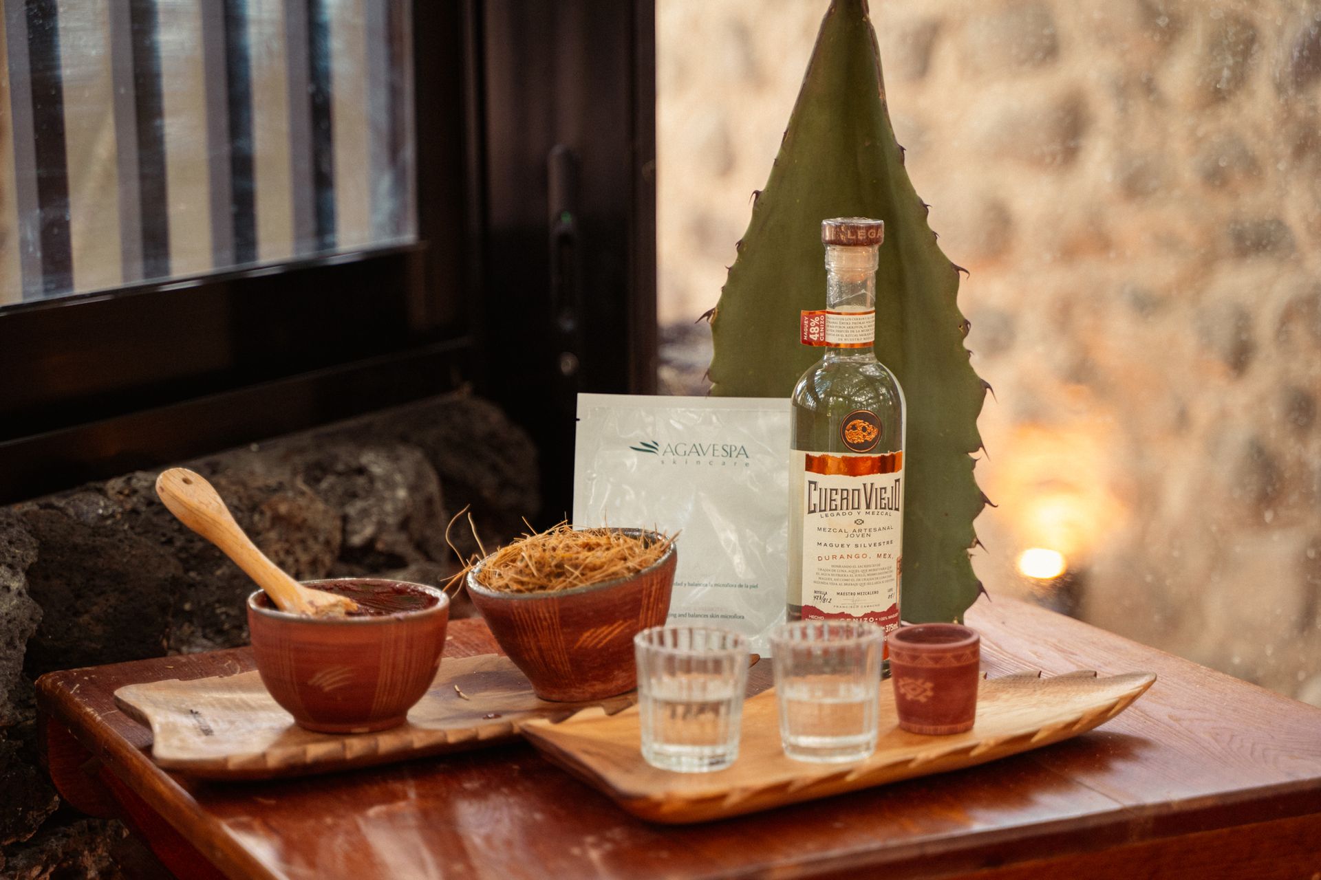 Juego de tequila con vasos de chupito, botella e ingredientes sobre una bandeja de madera en un ambiente rústico.