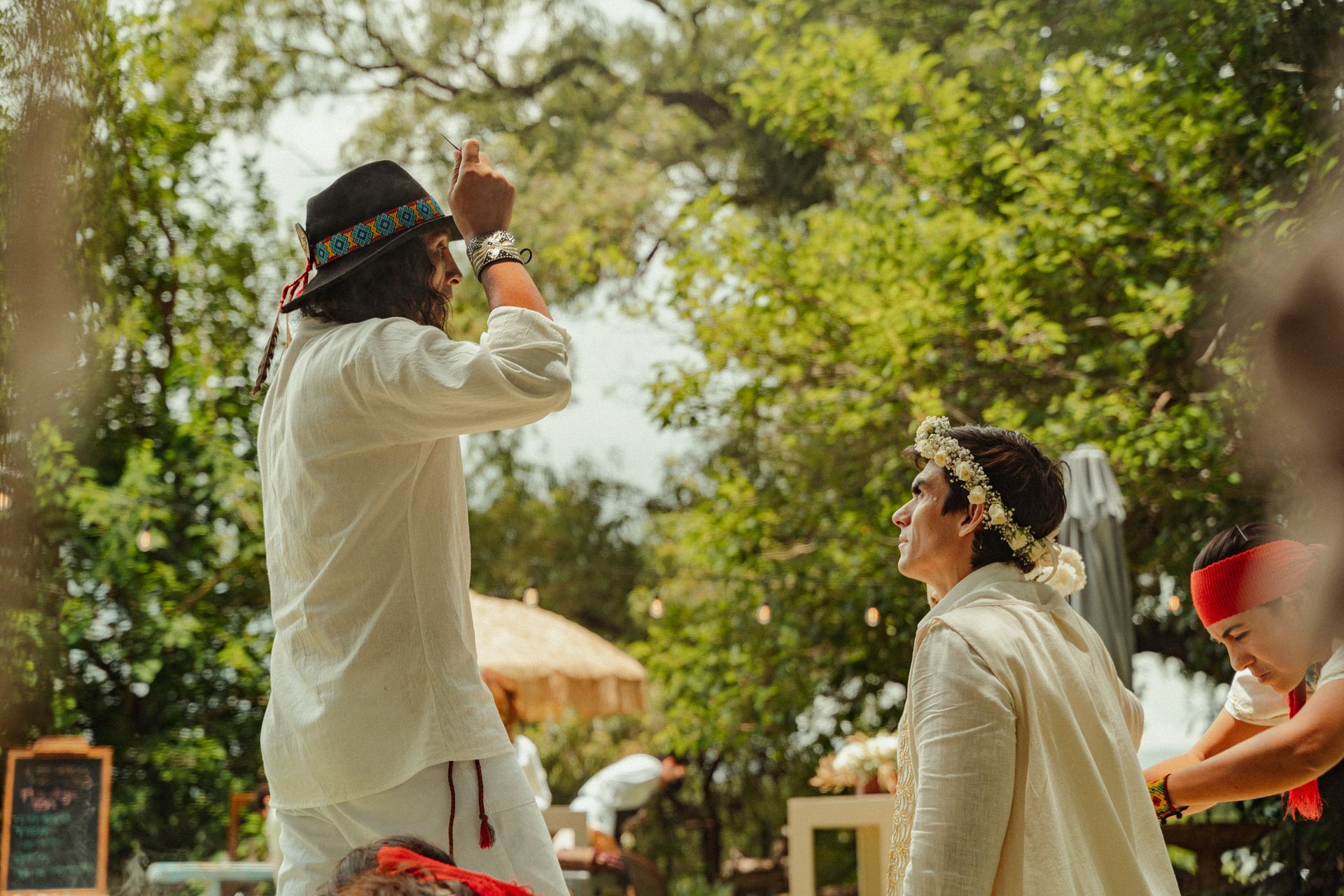Un hombre vestido de blanco realiza una ceremonia al aire libre para otro hombre que lleva una corona de flores.