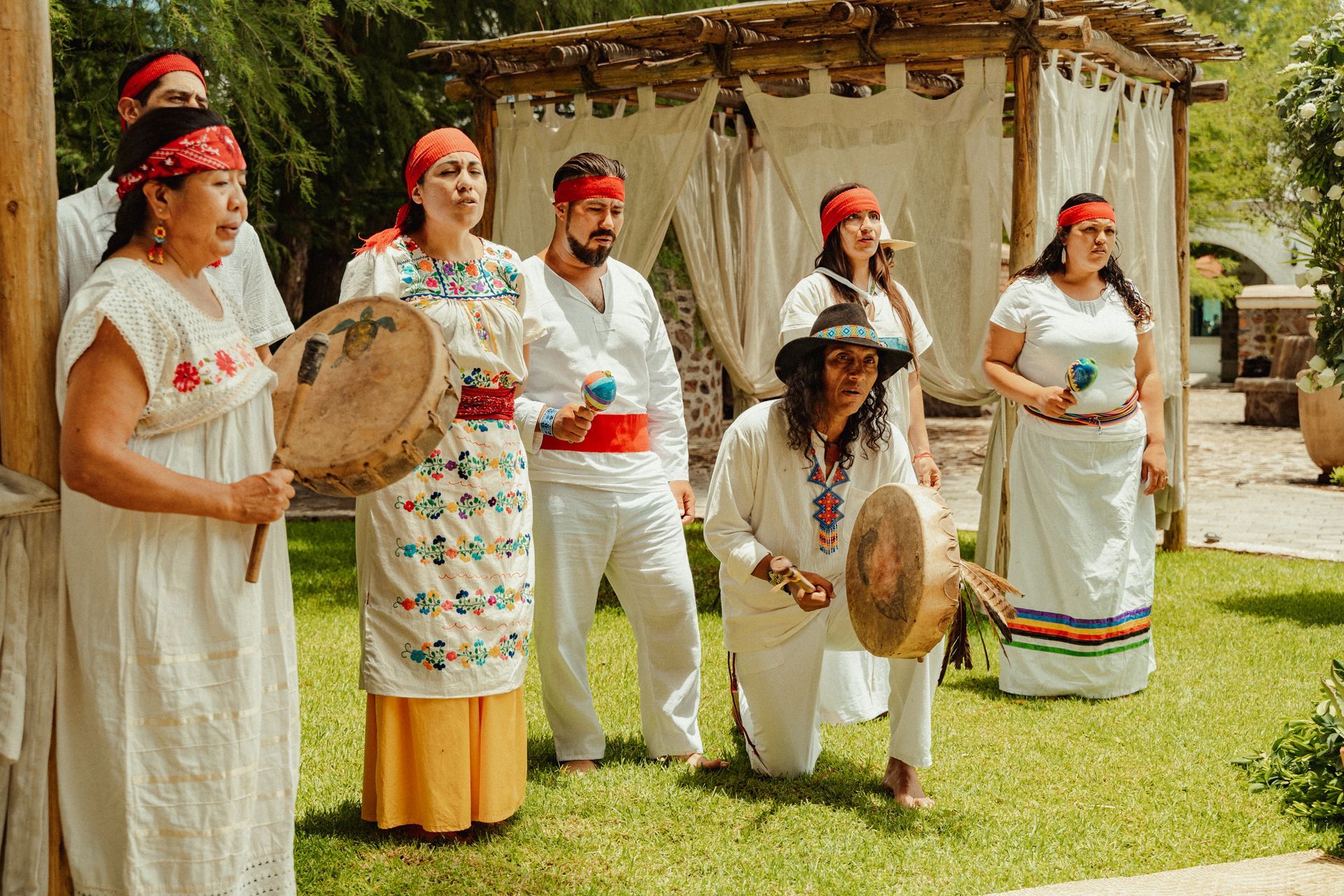 Grupo de personas vestidas de blanco y con cintas rojas en la cabeza, con tambores, al aire libre en un espacio verde.
