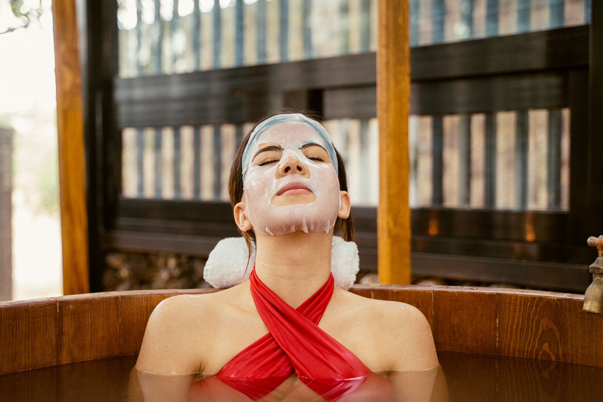Mujer con top rojo, mascarilla facial, relajándose en una bañera de madera con agua hasta el pecho.