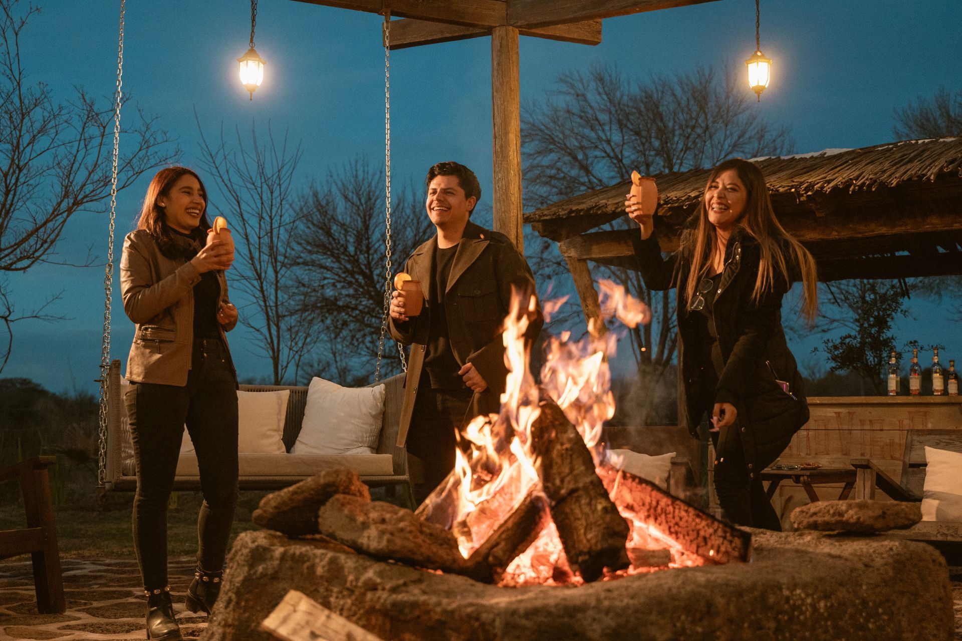 Three people gathered around a campfire at dusk. One gives a thumbs up; all hold food.