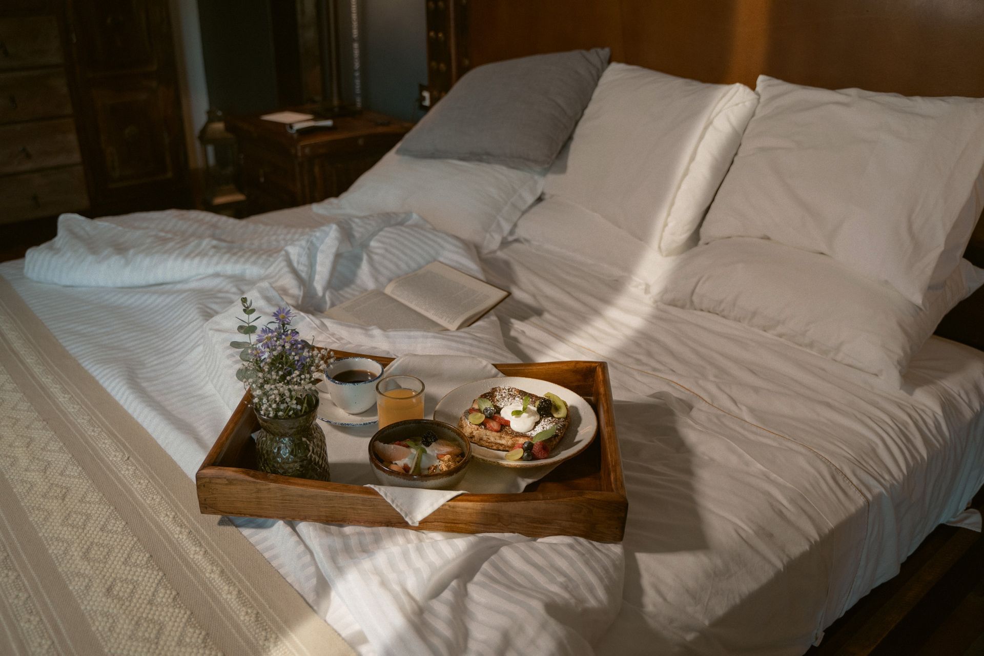 Bandeja de desayuno con comida, flores y un libro sobre una cama con sábanas blancas.