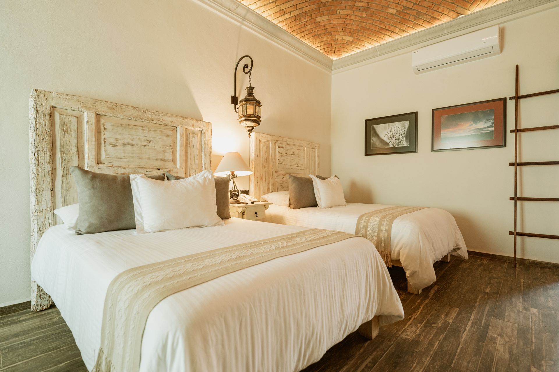 Dormitorio con dos camas, ropa de cama blanca, cabeceros decorativos, suelo de madera, farol, obras de arte y escalera.