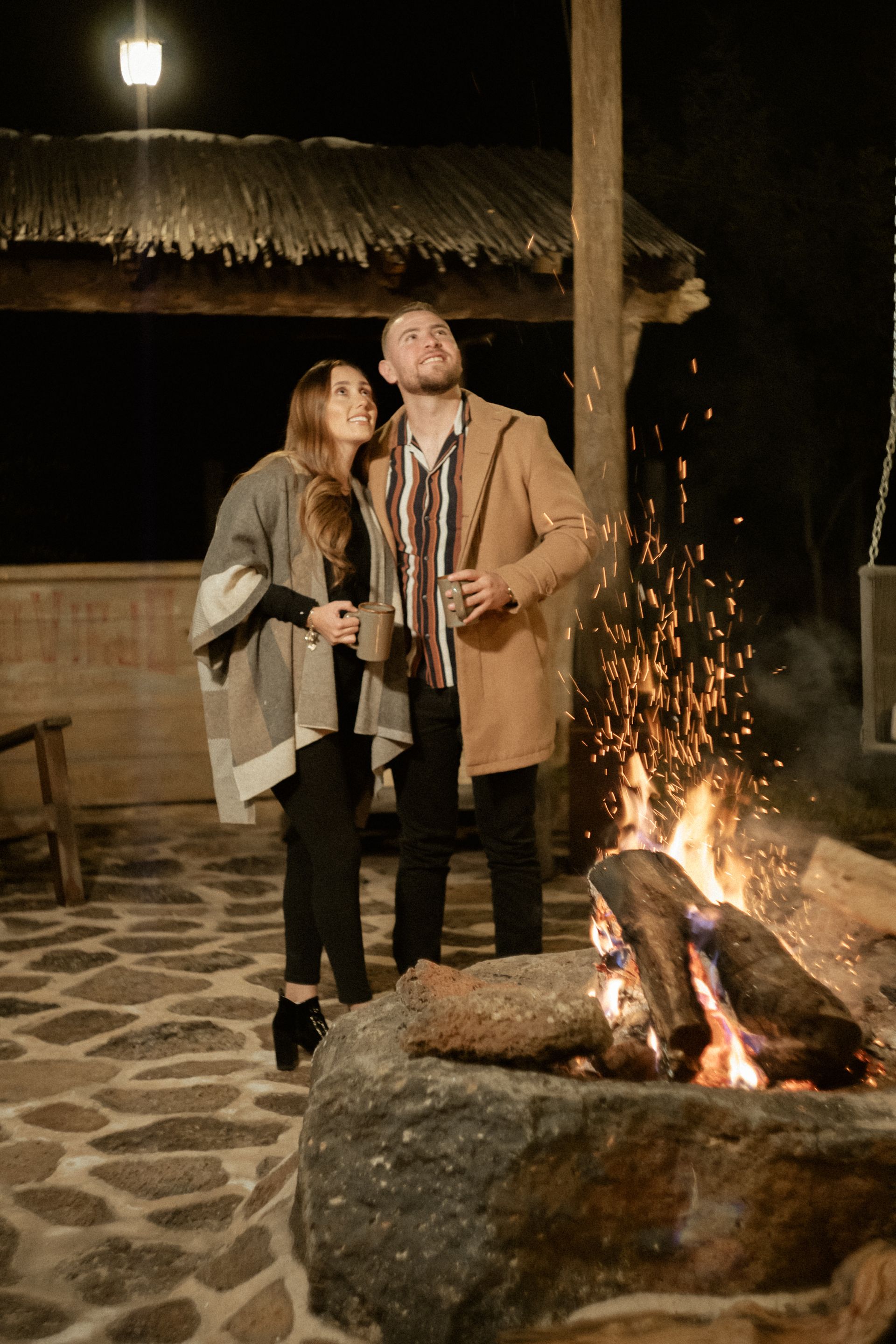 Pareja de pie junto a una hoguera por la noche, mirando hacia arriba. Saltan chispas bajo una estructura de madera.