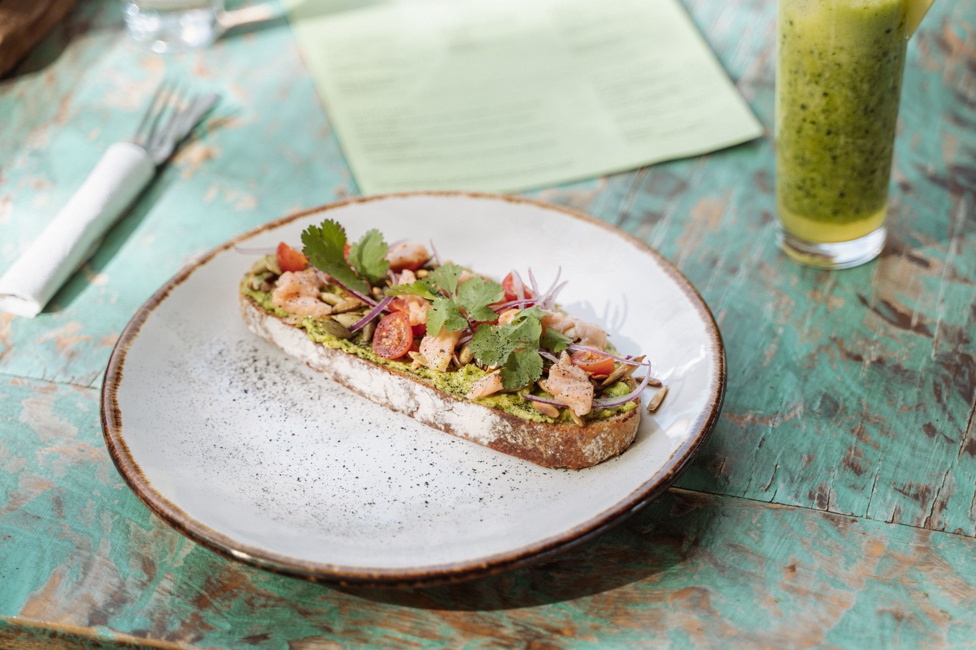 Tostada con salmón, aguacate y hierbas servida en un plato, acompañada de una bebida verde y cubiertos.