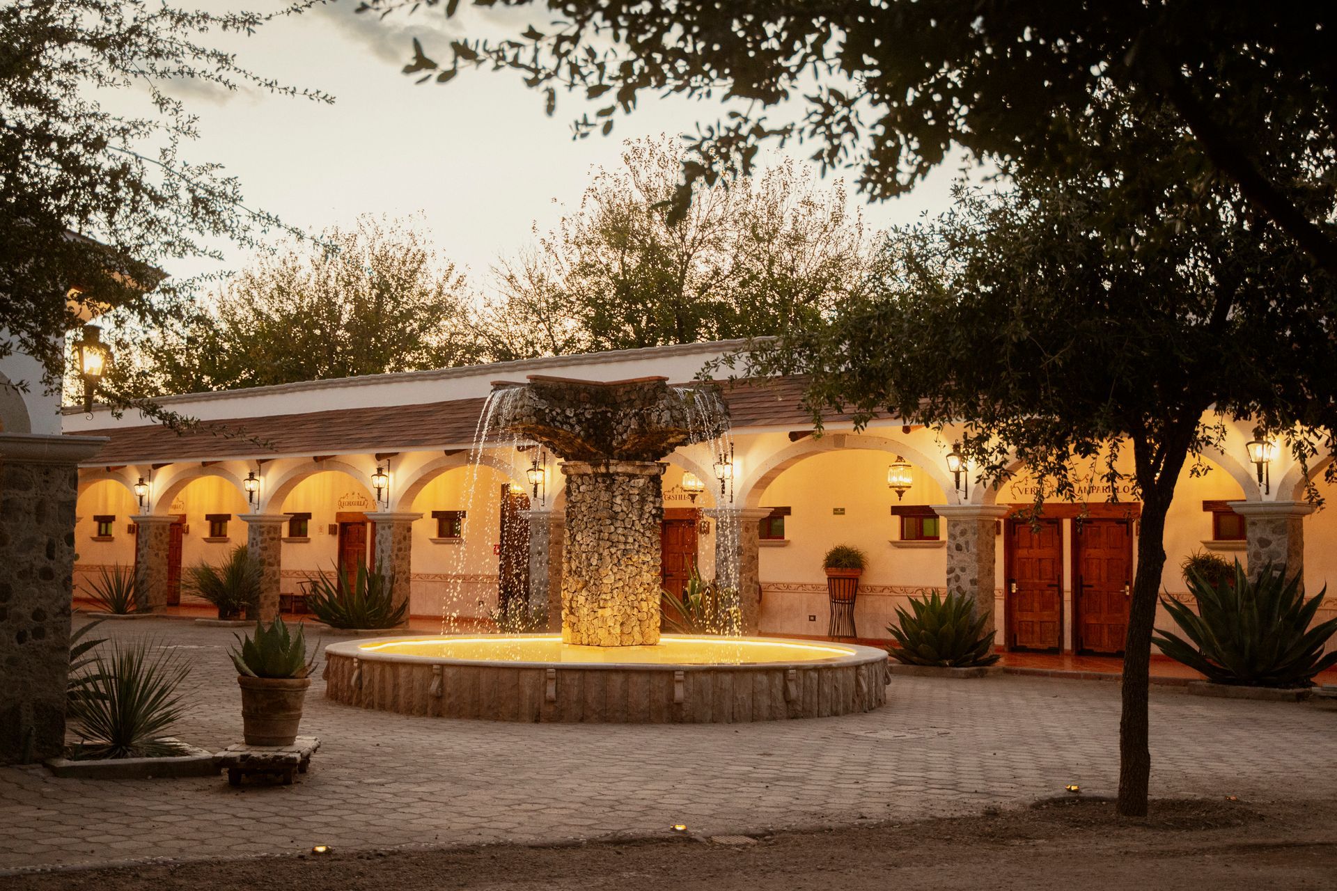 Fuente en el patio de un edificio con pasarelas arqueadas, árboles e iluminación cálida.