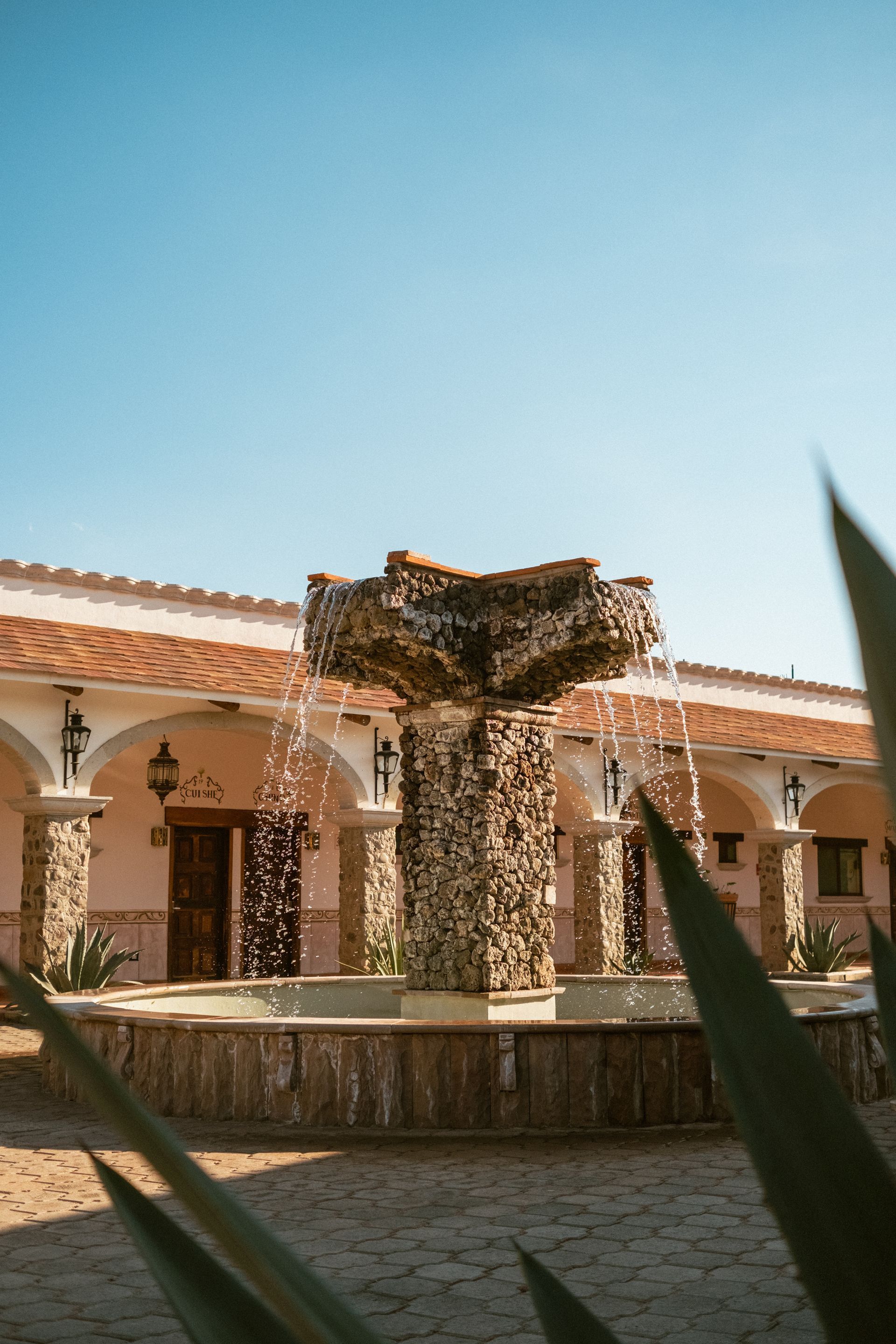 Fuente en el patio, de piedra, con agua que fluye. Edificio con arcos y tejado de tejas rojas, cielo azul.