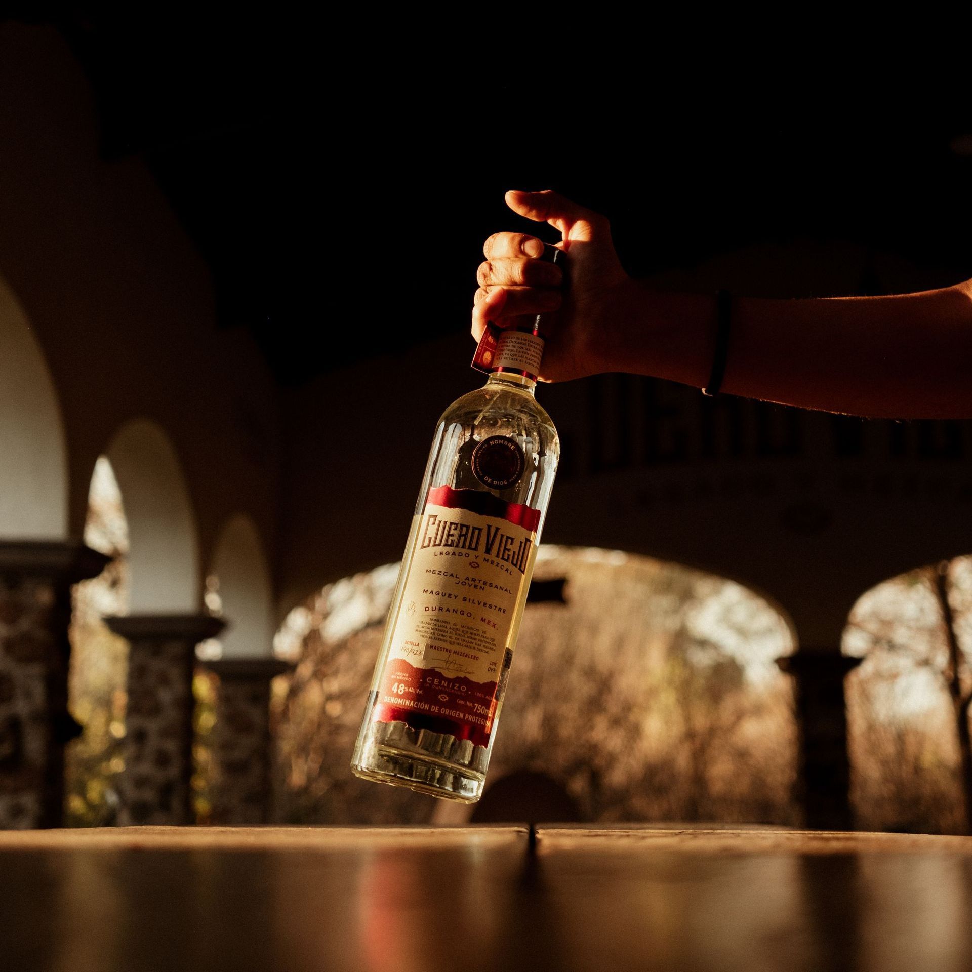 Hand holding a bottle of liquor, set in front of an outdoor structure with arches.