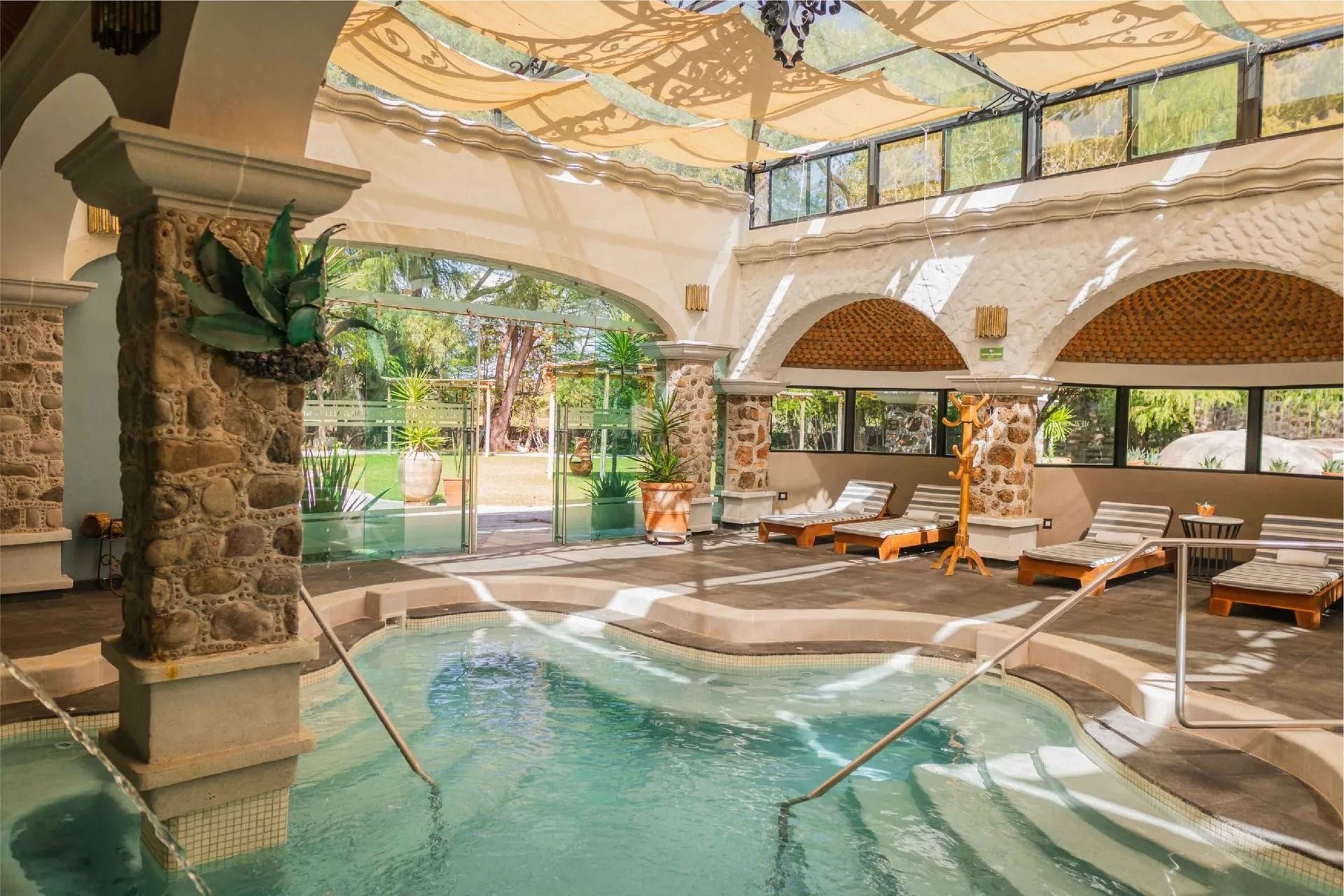 An indoor pool area with stone columns, arched ceilings, and lounge chairs arranged on a tile floor.