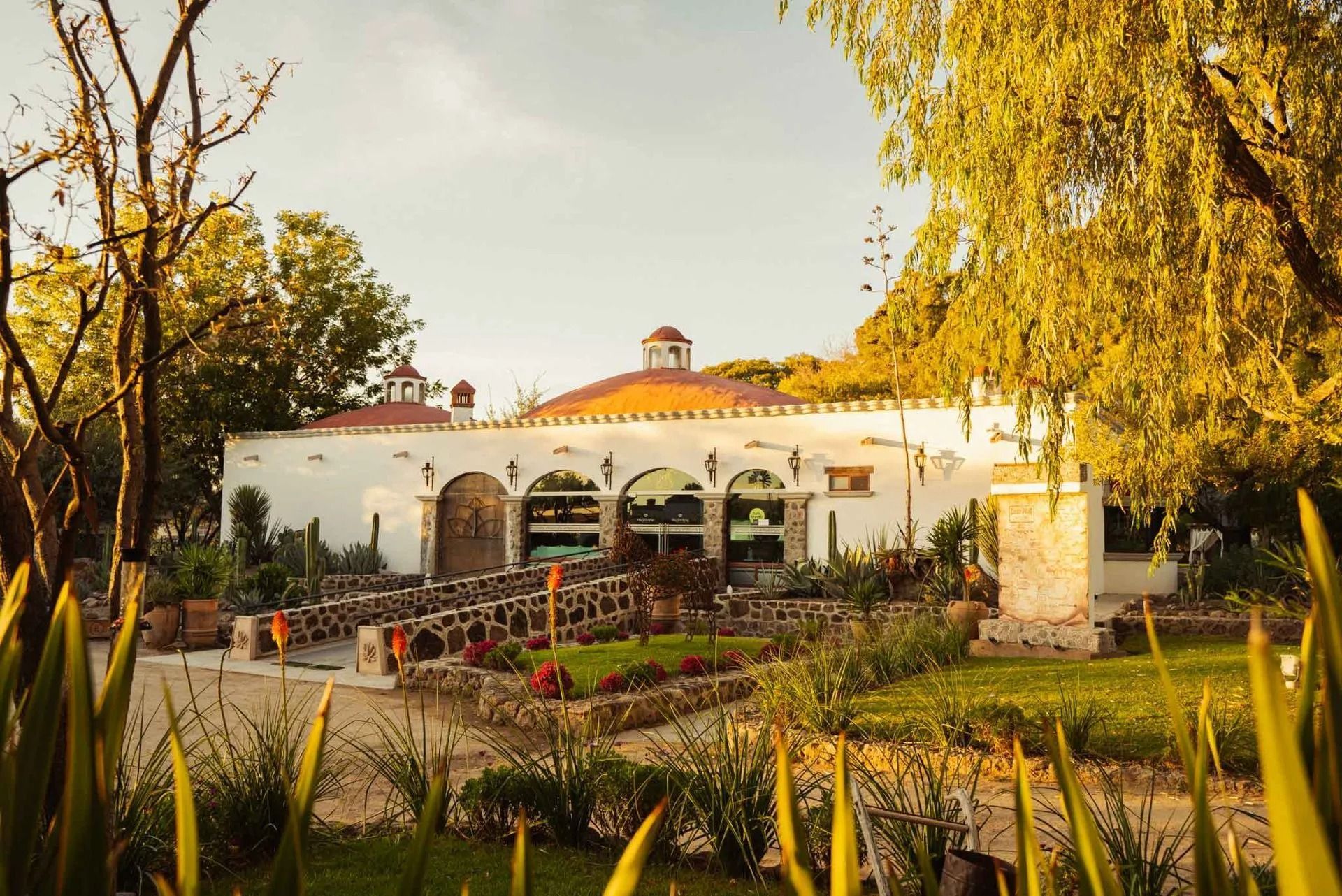 White hacienda-style building with arched entrances, red roofs, and surrounding greenery under warm, golden hour light.