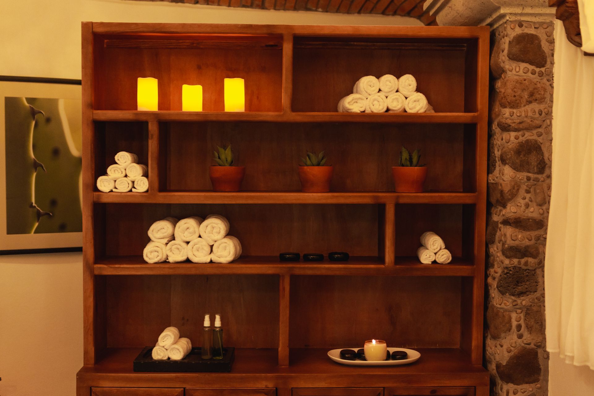 Estantería de madera con toallas, velas y plantas, junto a un pilar de piedra.