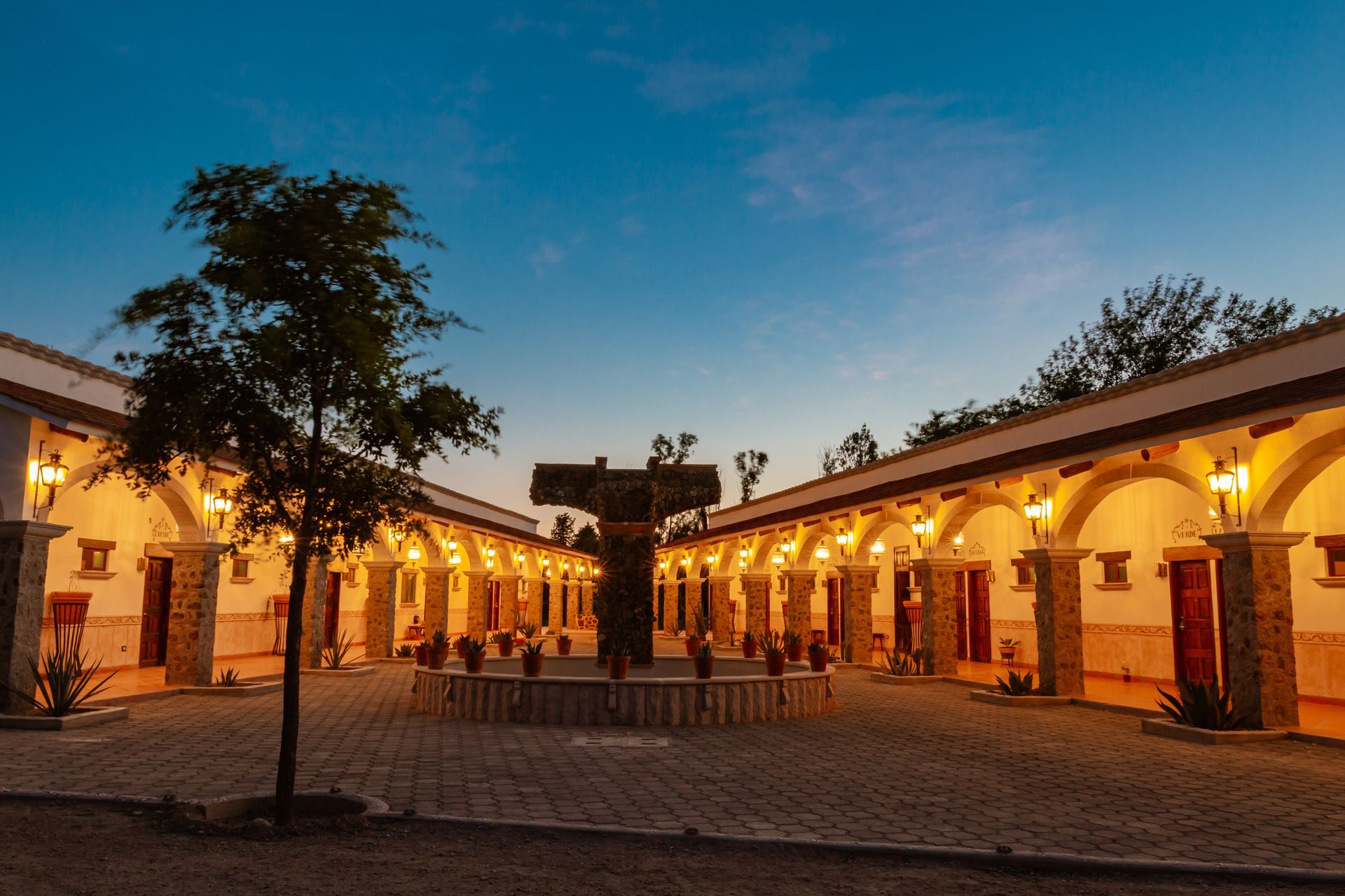 Courtyard with arched walkways, lit by warm lights. A stone cross fountain is centered. Dusk sky.