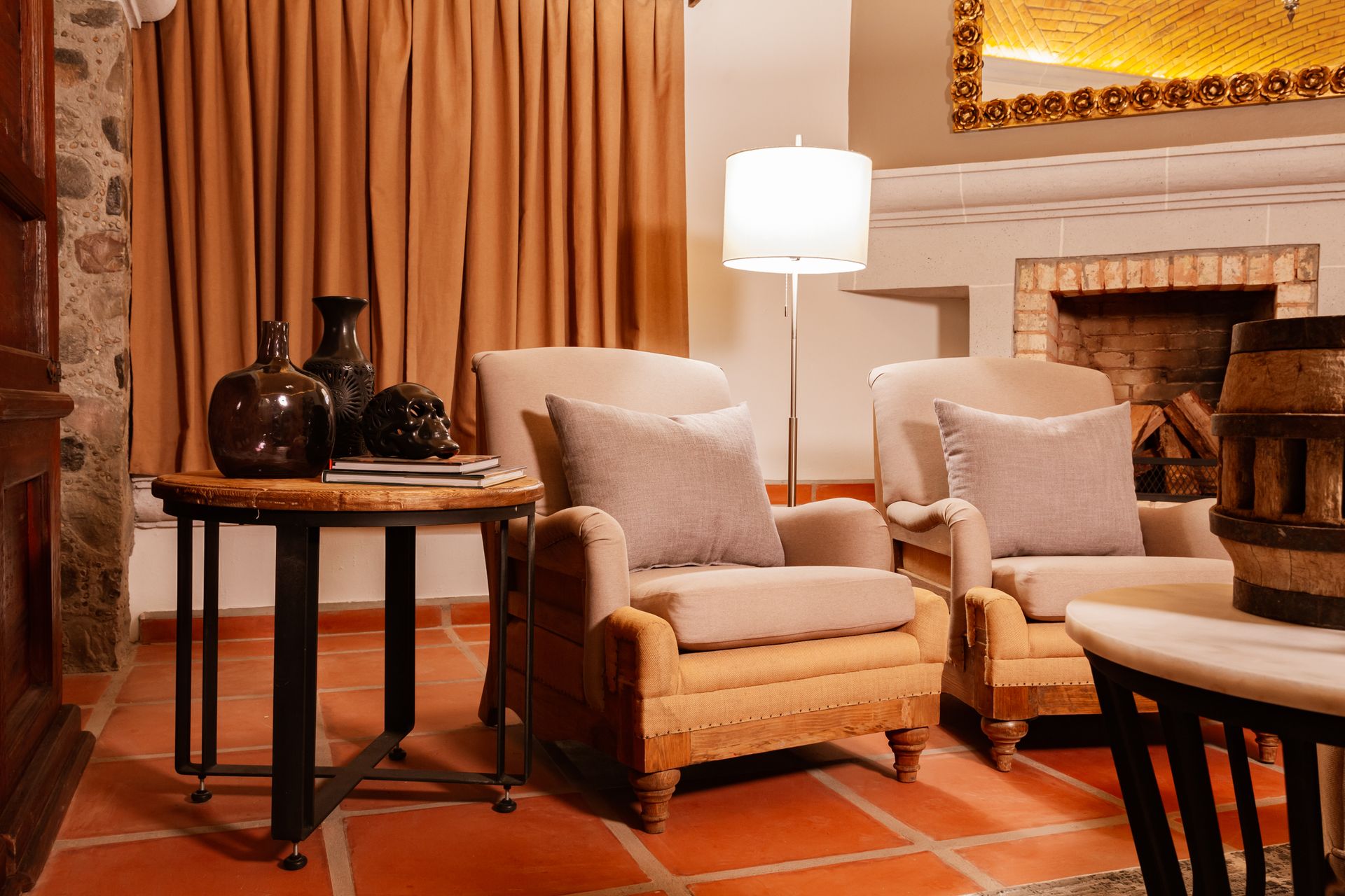 Cozy living room with armchairs, a side table with decor, and a fireplace. Orange tiled floor, brown curtains.