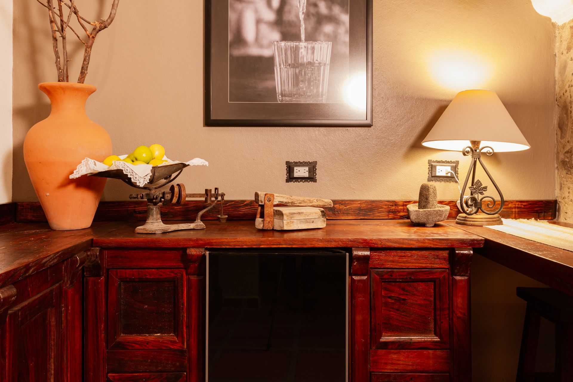 Warm-toned wooden bar with a vase, fruit bowl, lamp, and framed art.