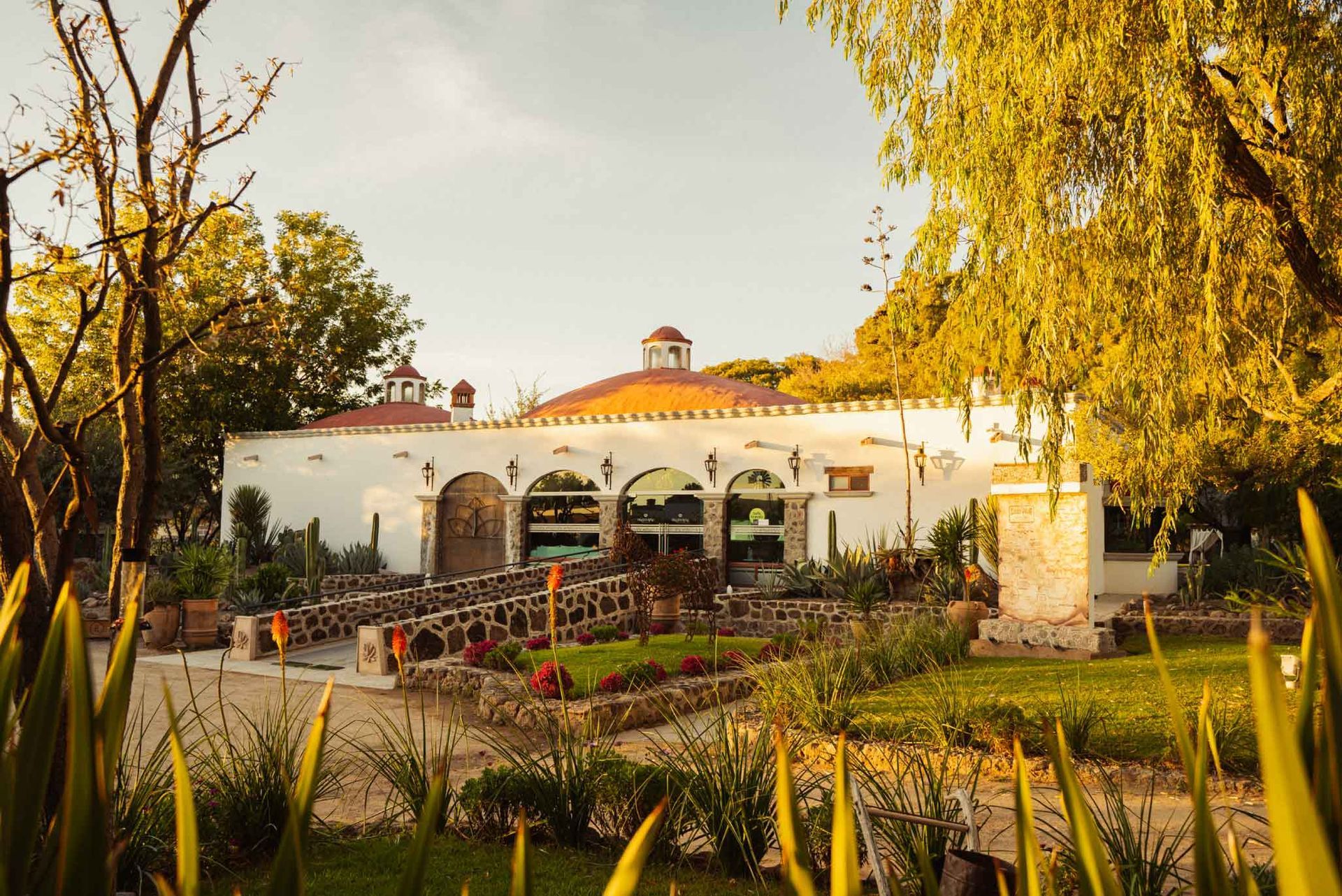 White building with arched windows and orange roof in a garden setting, illuminated by sunlight.