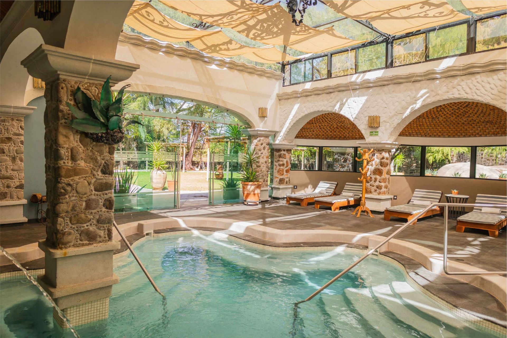 Indoor spa with pool, lounge chairs, and arched doorways leading to outdoor greenery.
