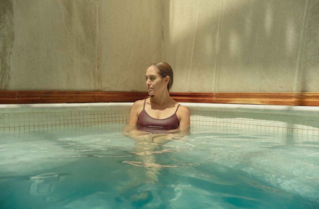 Mujer en traje de baño morado relajándose en un jacuzzi turquesa, mirando hacia otro lado. Interior, paredes alicatadas.