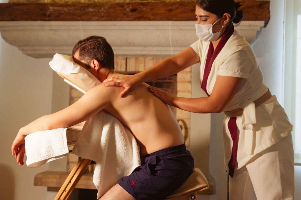 Persona recibiendo un masaje sentada de un terapeuta con mascarilla en interiores.