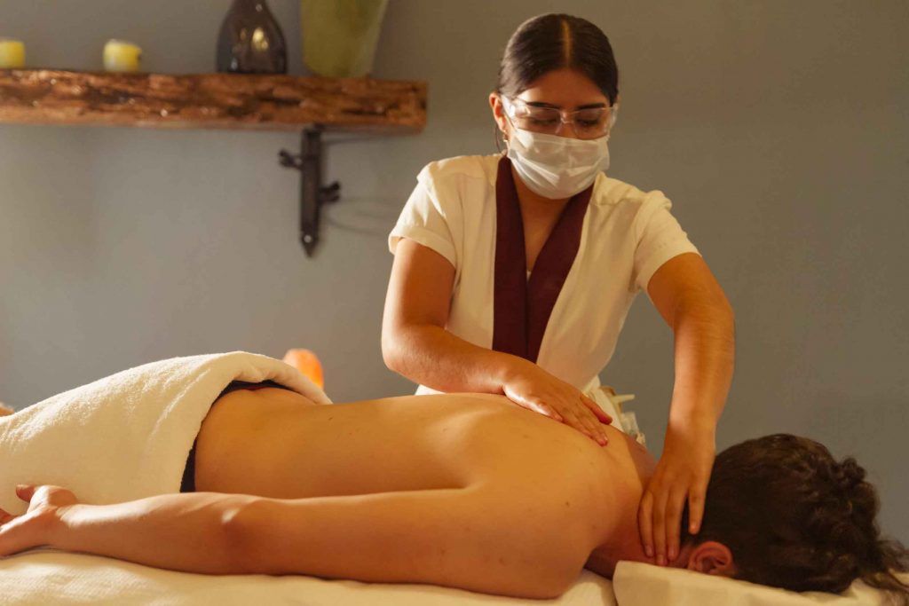 Persona recibiendo un masaje de espalda en un spa. Terapeuta con uniforme blanco y mascarilla.