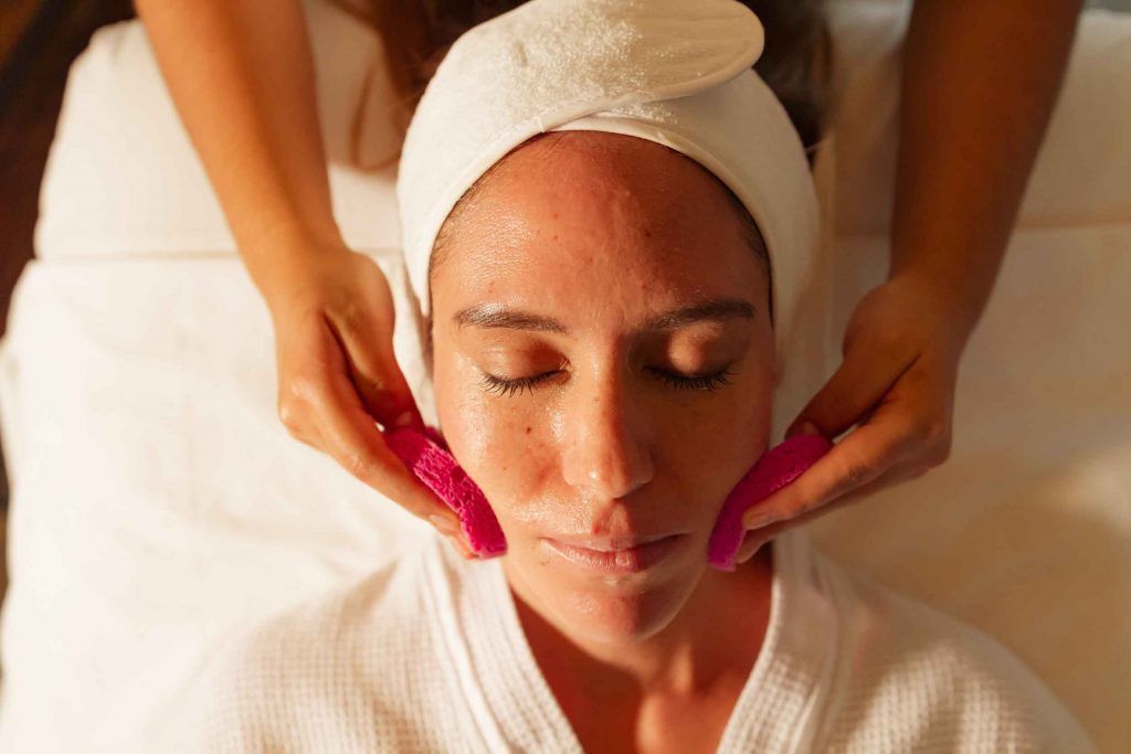 Mujer recibiendo un masaje facial en un spa. Sus manos sostienen almohadillas rosas sobre sus mejillas.