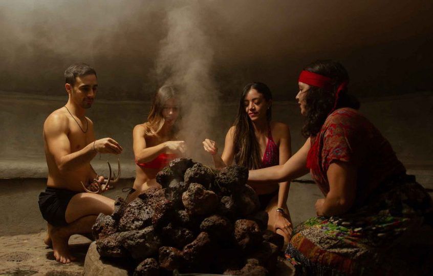Cuatro personas en un espacio cerrado y con poca luz, alrededor de una pila de piedras calientes, del que se eleva vapor.