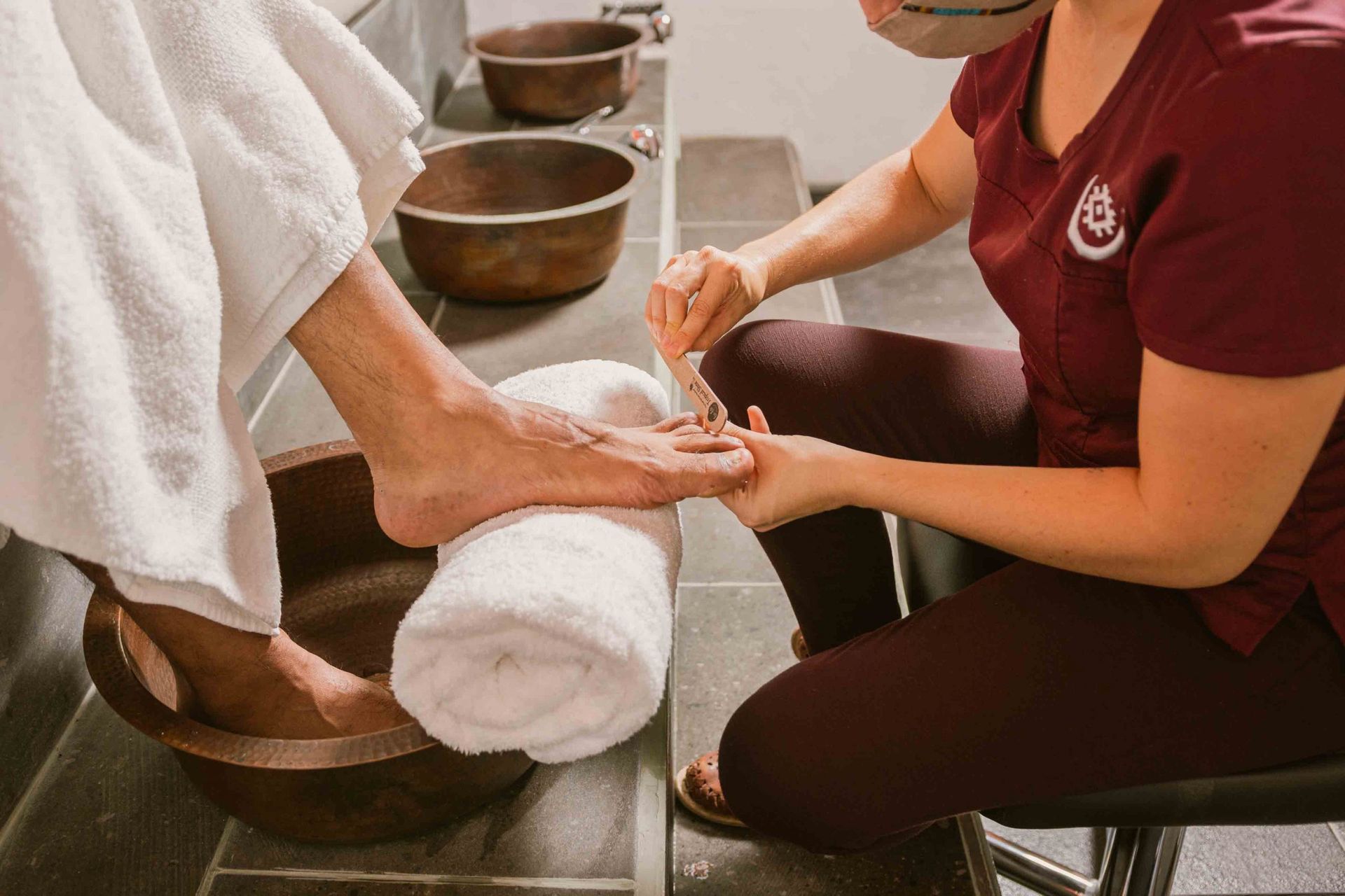 Persona recibiendo una pedicura; técnico limando las uñas de los pies. Cuencos de cobre, toallas blancas.