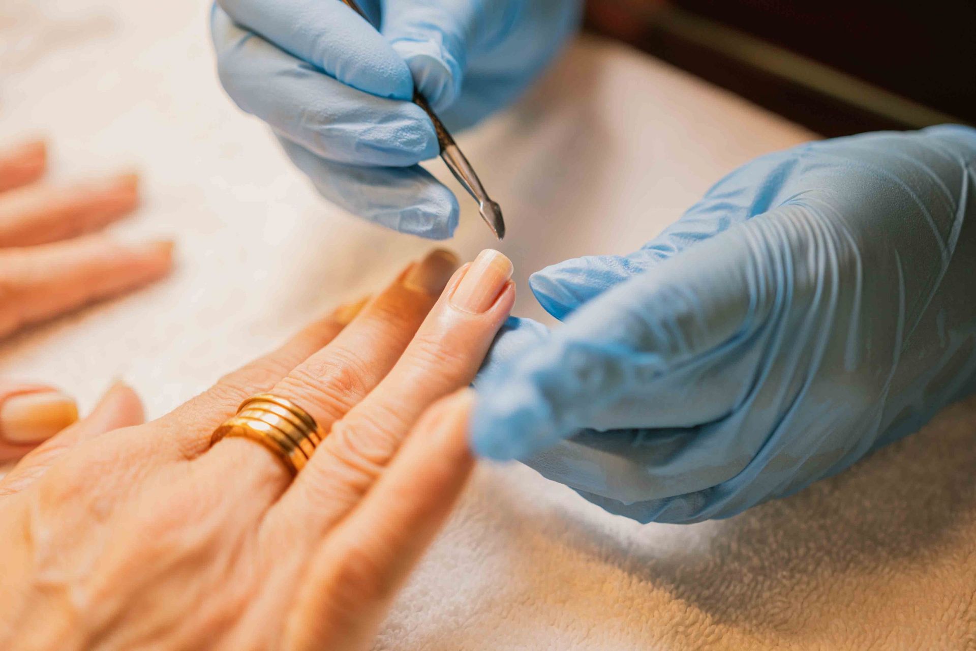 Manicurista utilizando una herramienta en la uña; manos enguantadas, anillo de oro, piel clara.