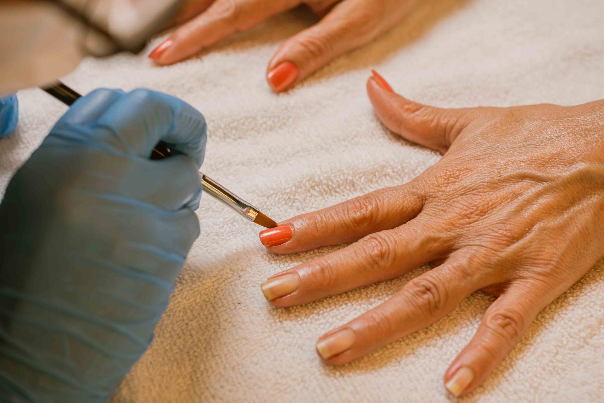 Manicurista aplicando esmalte de uñas color coral en la mano de una clienta, primer plano. Guante azul, toalla blanca.