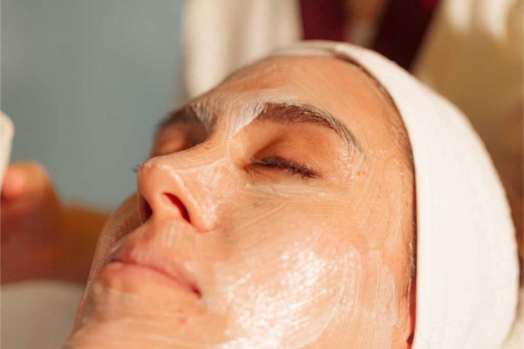 Persona con los ojos cerrados, recibiendo un tratamiento facial. Rostro cubierto con crema blanca.