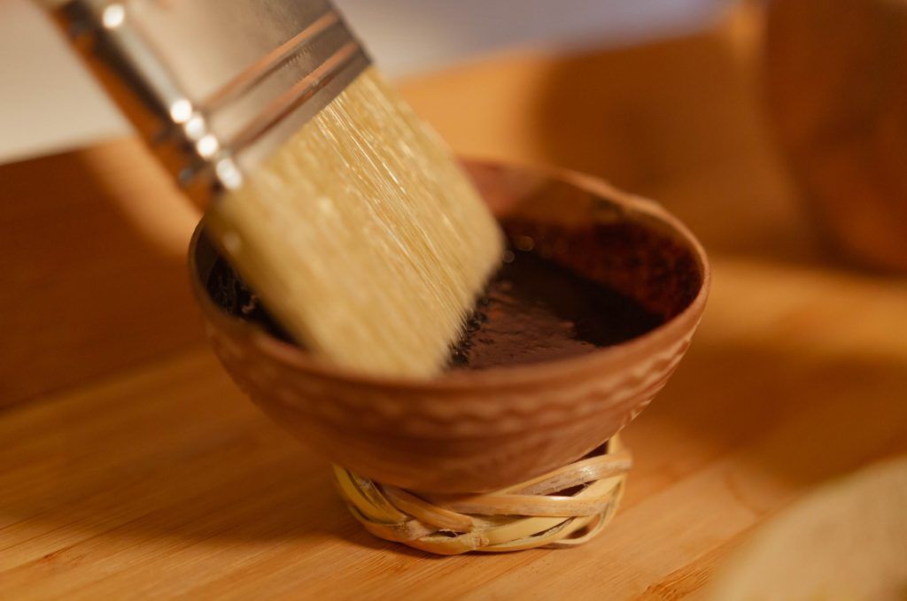 Un pincel sumergido en un líquido oscuro dentro de un pequeño cuenco marrón, colocado sobre una superficie de madera.