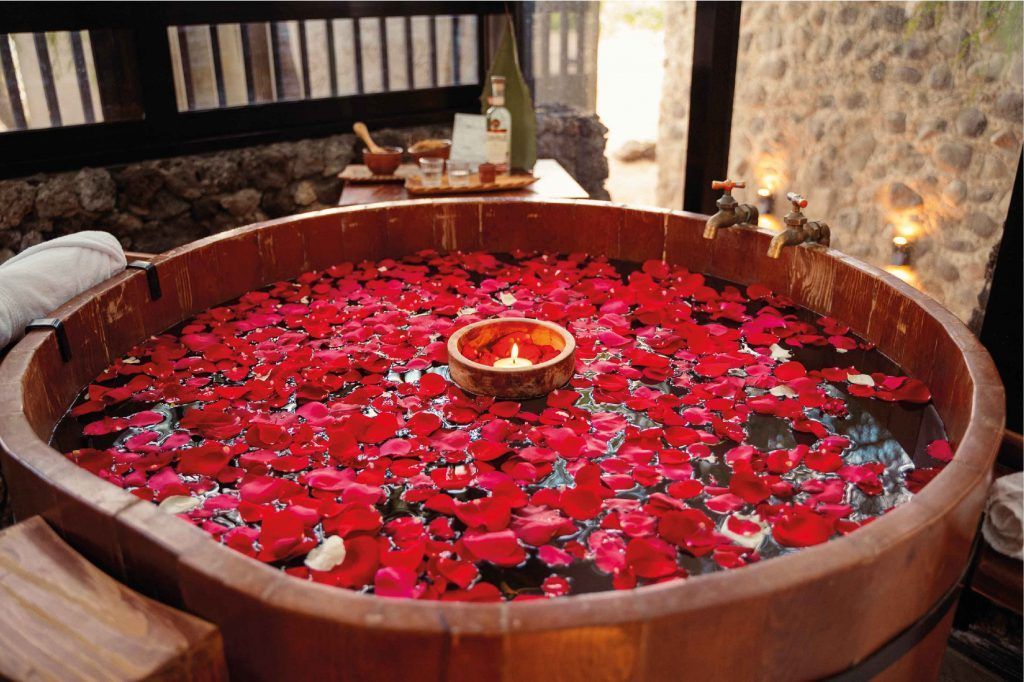 Bañera de madera llena de pétalos de rosa roja, vela encendida y ambiente de spa.