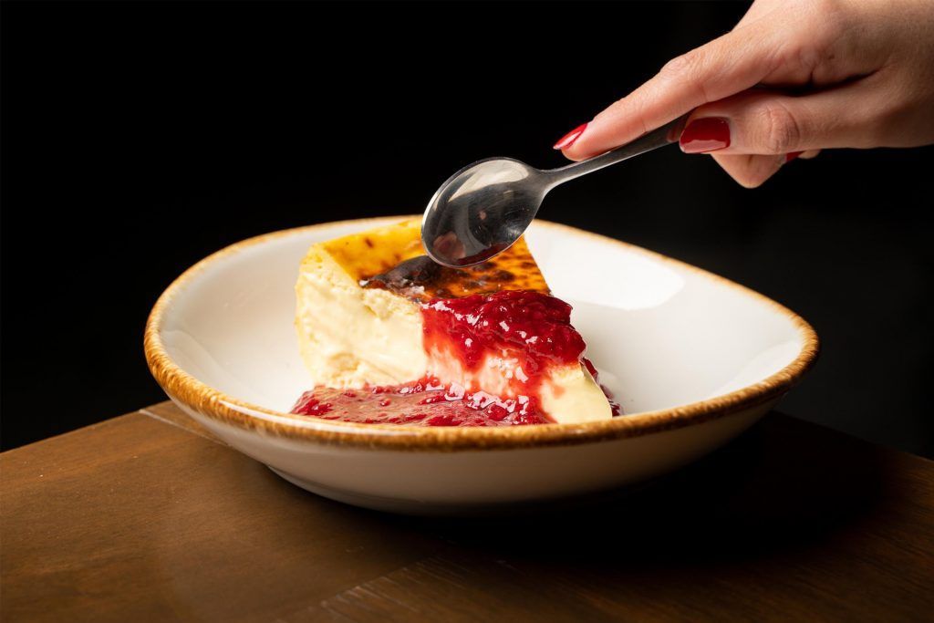 Slice of cheesecake with berry sauce in a bowl, hand holding spoon.