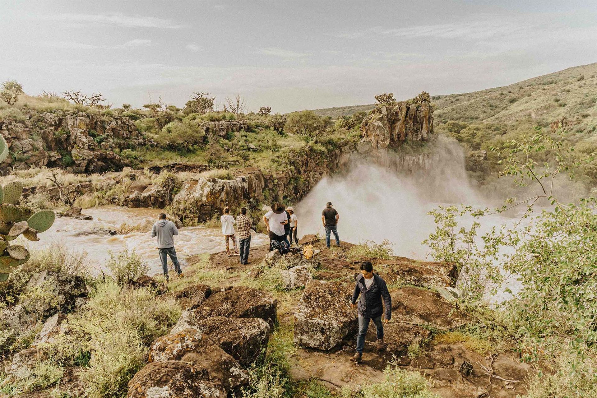 People watch a geyser erupt in a rocky, green landscape.