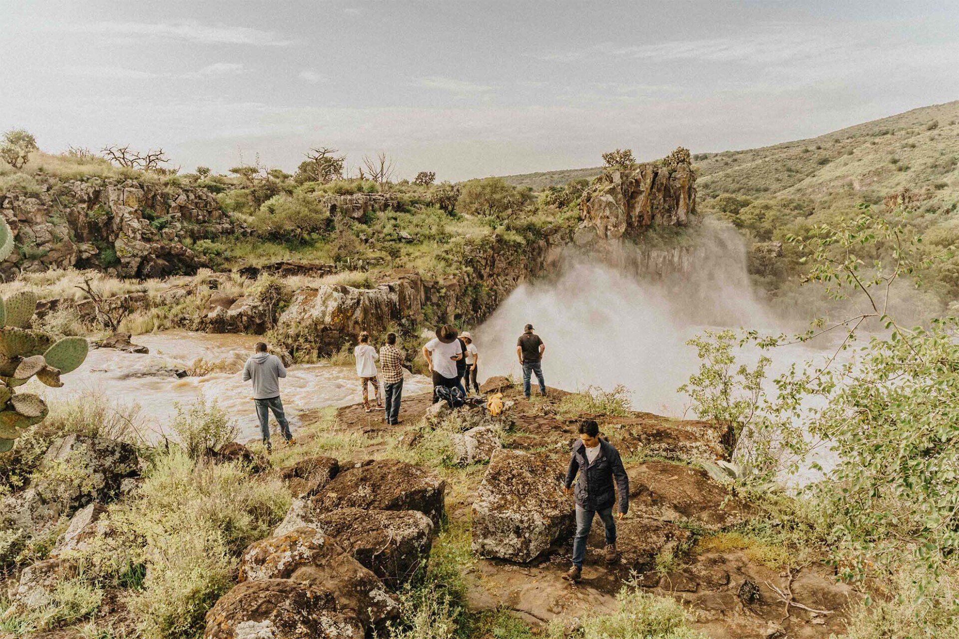 People near a waterfall, standing on rocks and taking photos. Green and brown landscape.