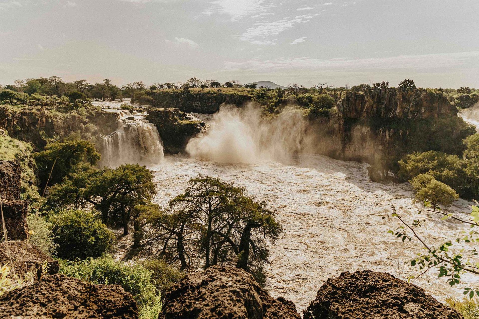 Waterfalls cascading down cliffs, creating a foamy river surrounded by trees, under a sunny sky.