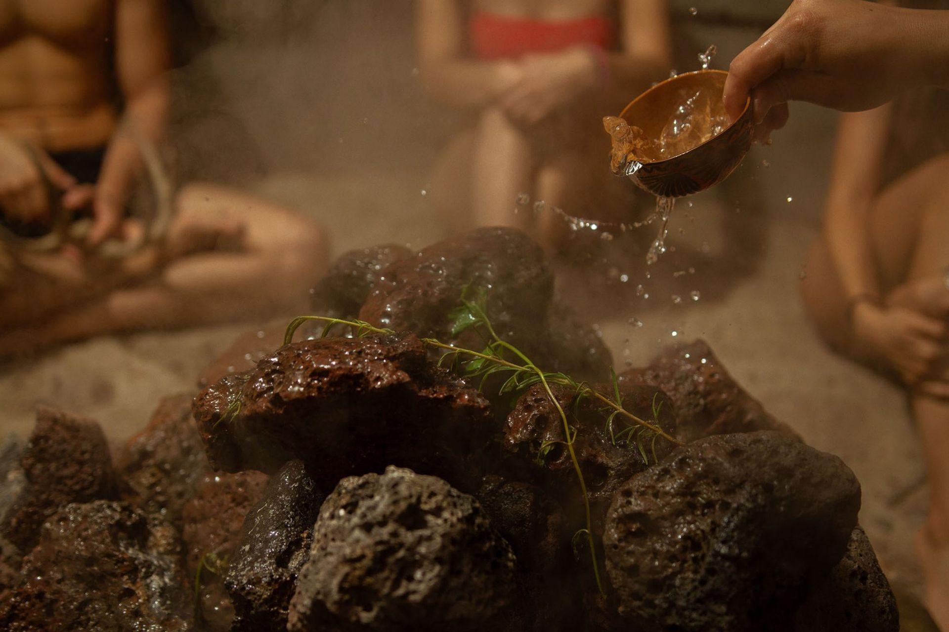 Person pours water from a wooden bowl onto a hot rock in a sauna, creating steam.