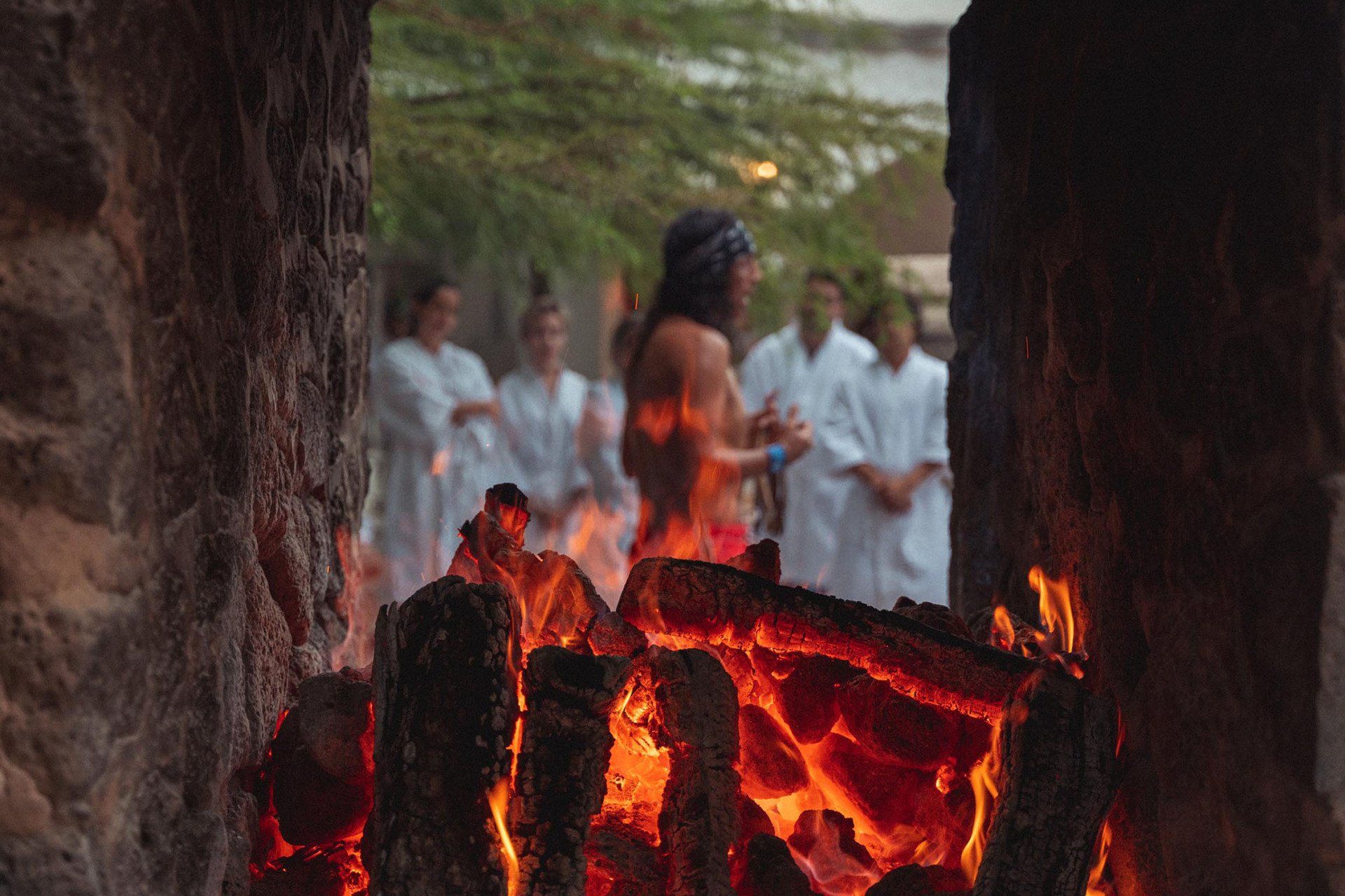 Fire pit with burning wood; people in white robes stand behind a man, who is shirtless, in front of the flames.