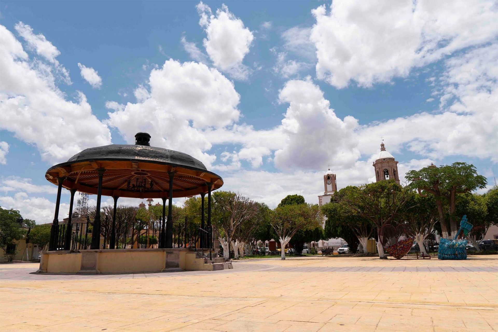 A gazebo sits in a town square with a church in the background under a blue sky with clouds.