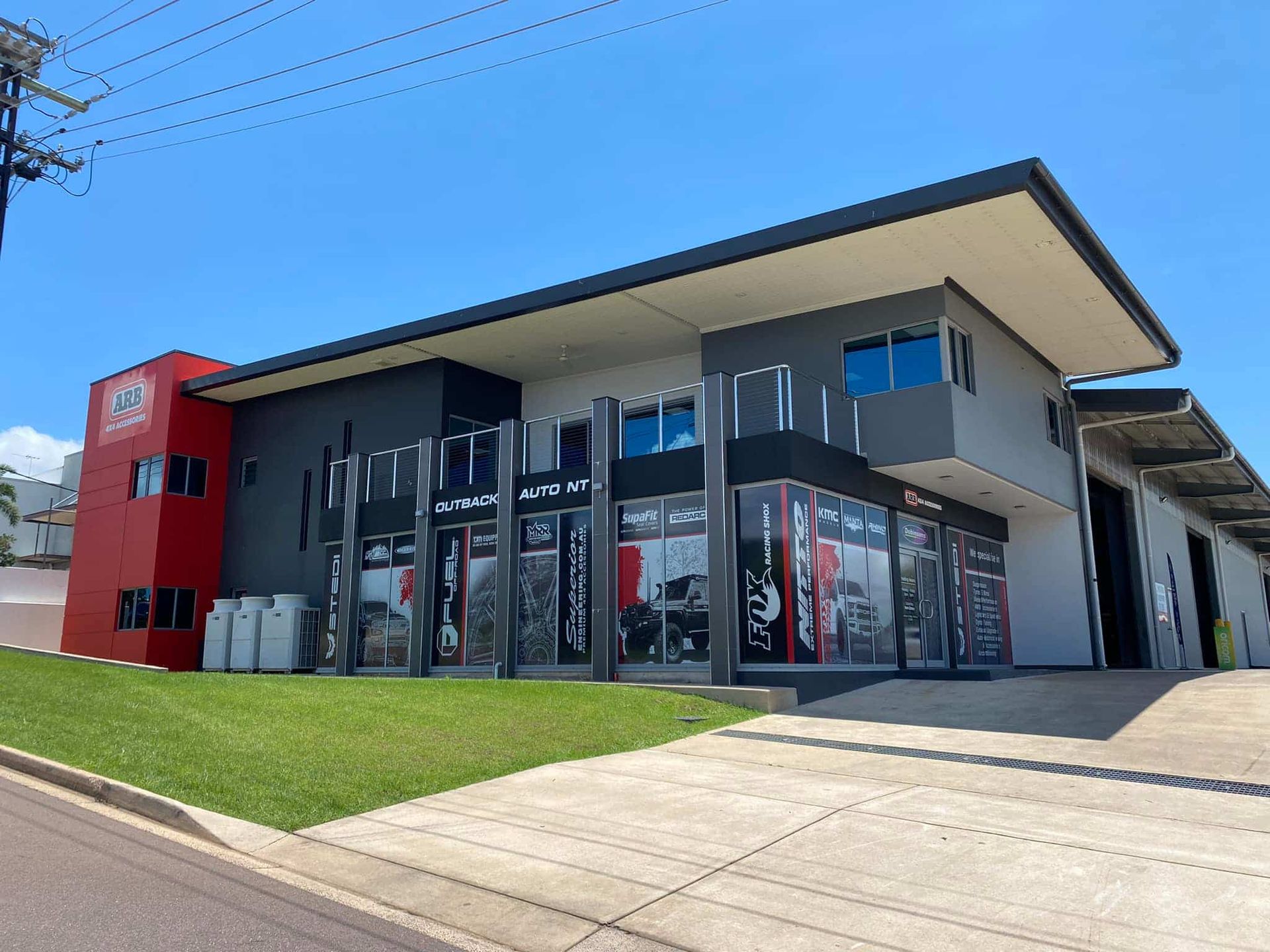 Modern glass-front commercial building with a red side wall and sloped driveway under a blue sky — Outback Auto NT in Berrimah, NT