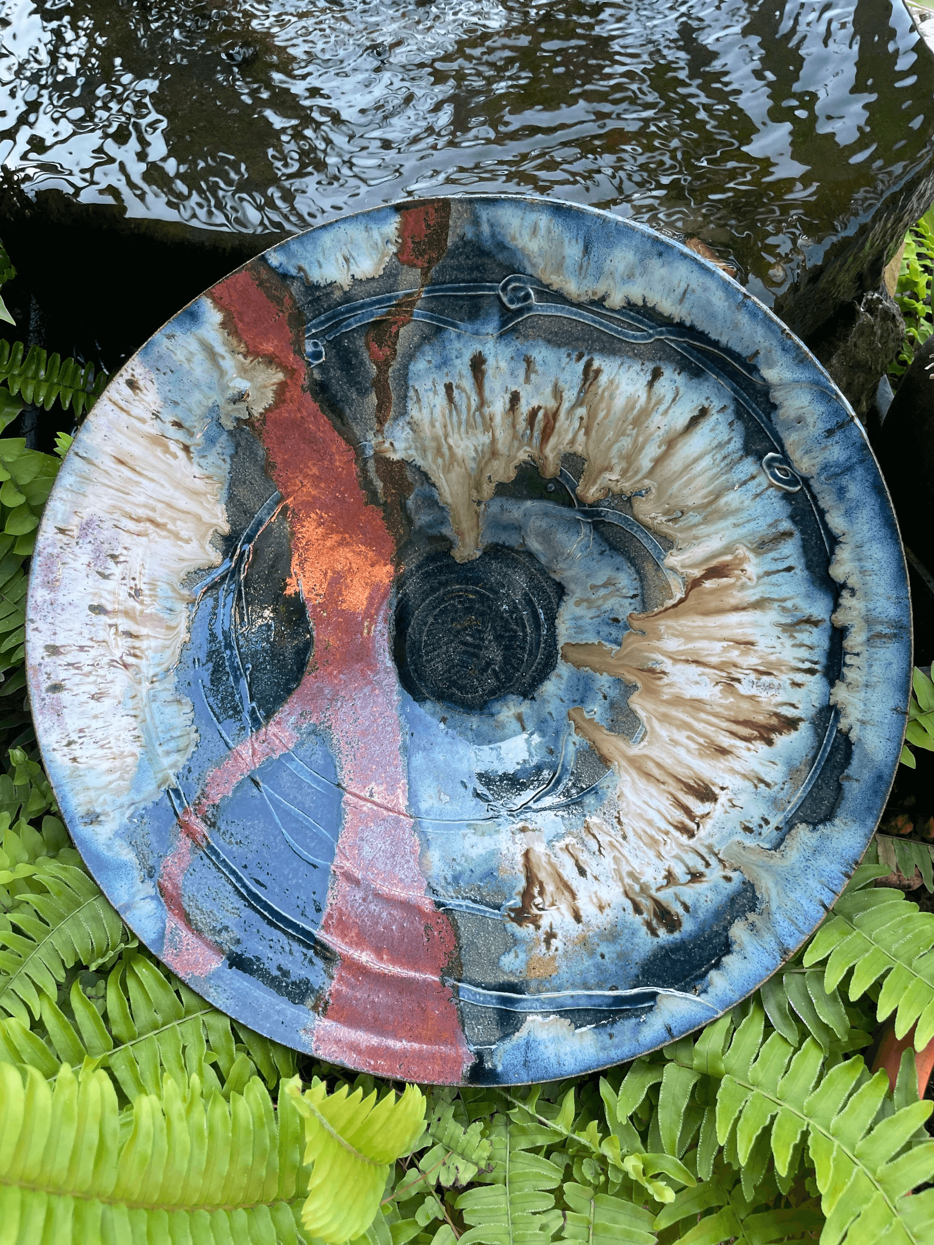 a large red, gold, blue, and black serving platter surrounded by ferns and a water feature